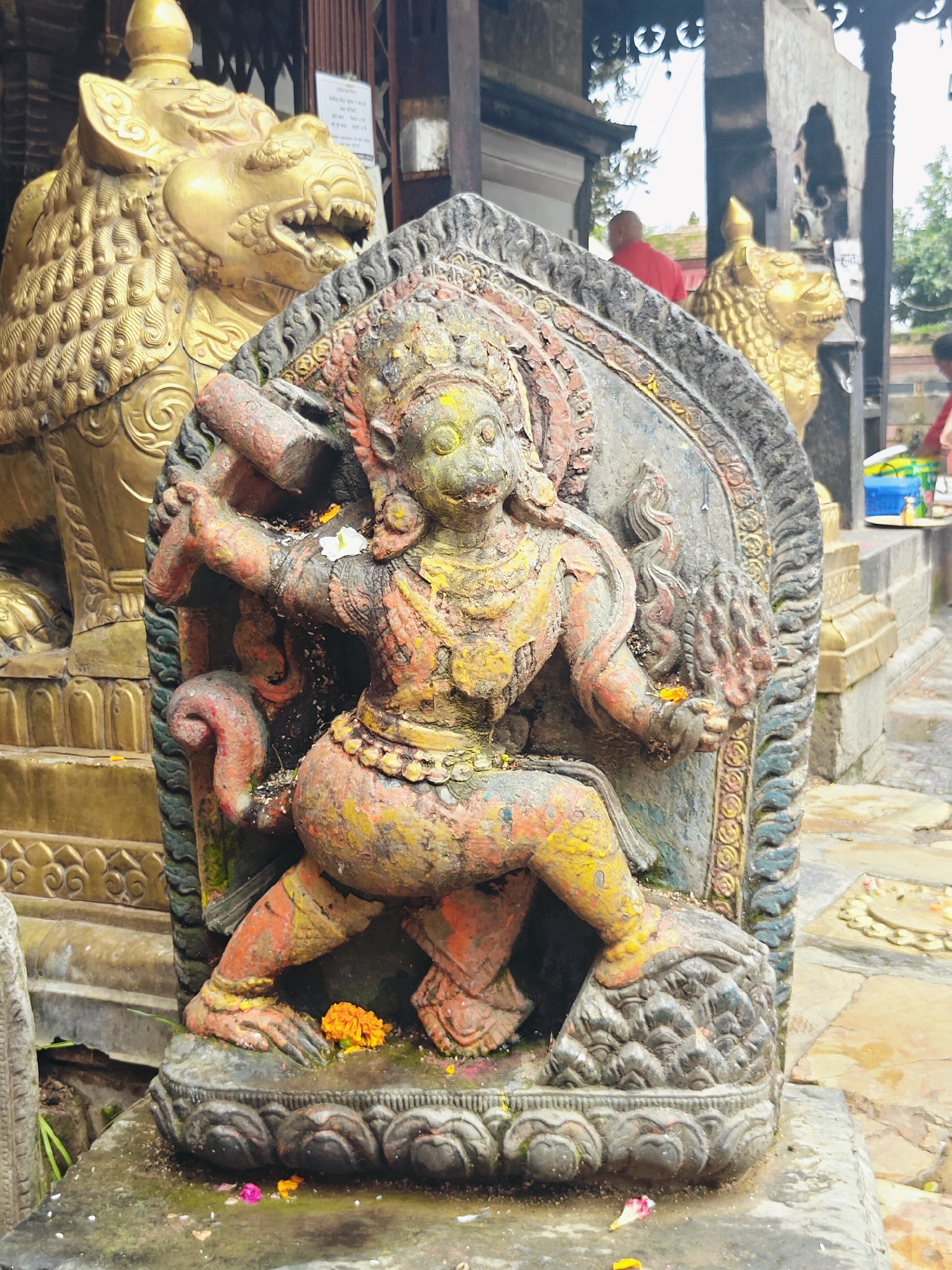 The image shows a weathered stone sculpture of a multi-armed deity, possibly a depiction of a wrathful aspect of a Hindu or Buddhist deity, in Kirtipur, Nepal. The sculpture is painted in faded colors of orange, yellow, green, and red, with visible wear and tear, suggesting age and exposure to the elements. The deity is depicted in a dynamic pose, with one leg bent and the other extended, and appears to be holding objects in its hands, though their exact nature is difficult to discern due to the damage. In the background, a large golden lion statue is visible, likely a guardian figure common in temples and sacred sites. The setting appears to be an outdoor courtyard or temple complex, with stone paving and architectural elements suggesting a religious or historical location. Several small flowers, likely offerings, are scattered around the base of the sculpture.
