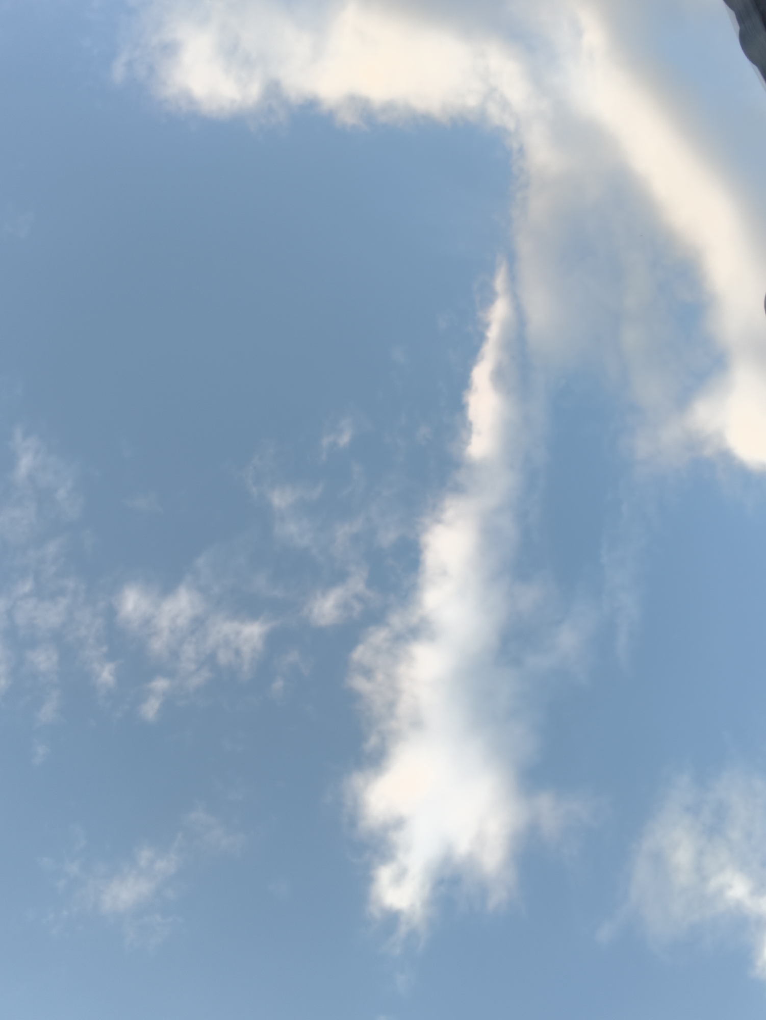 The image shows a partly cloudy blue sky. The clouds are white and wispy, with some areas appearing brighter than others, suggesting sunlight filtering through. The scene is captured from a low angle, looking upwards. There are no discernible landmarks, buildings, or people visible, making it impossible to determine the specific location within Chișinău, Moldova, or any other city. The focus is entirely on the sky and the cloud formations.