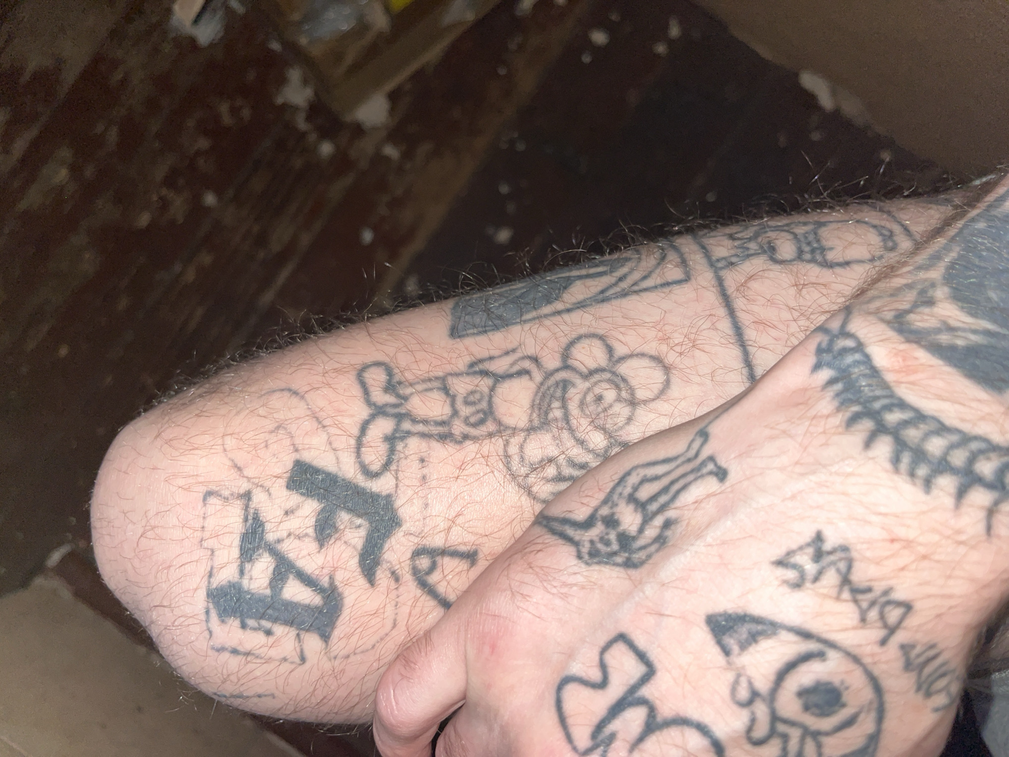 A close-up, high-angle shot shows a person's tattooed legs in an indoor setting with a wooden floor and dark background. The tattoos are black and varied in style, including lettering, cartoonish figures, and abstract designs. The skin appears pale and is covered in fine hairs. The lighting is dim, casting shadows and highlighting the texture of the skin and tattoos.