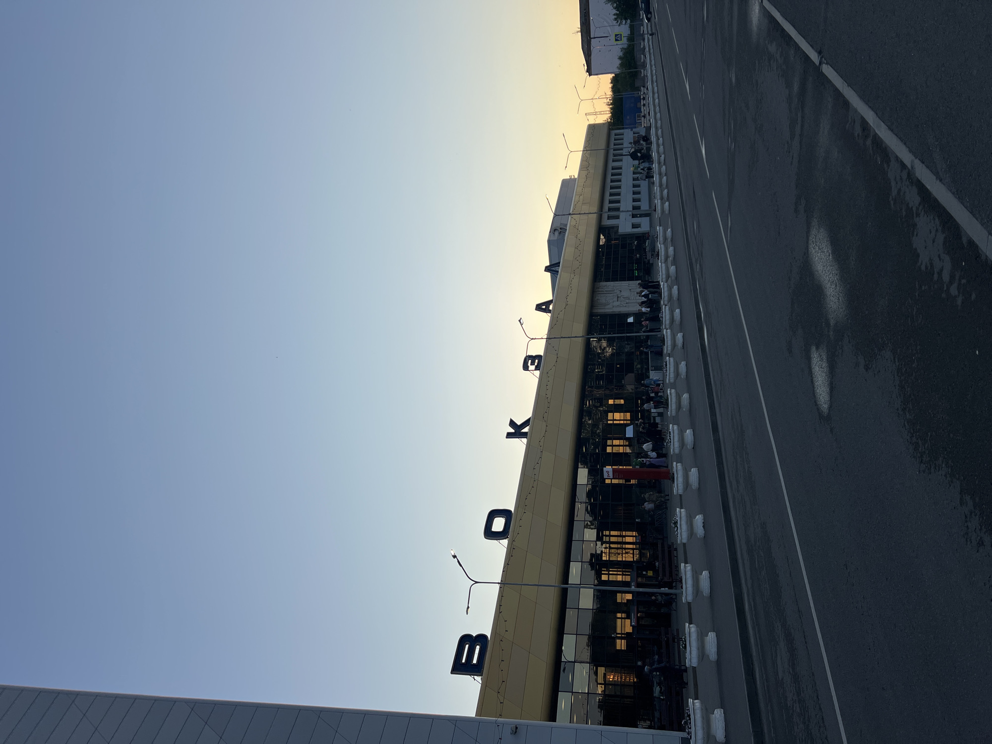 The image depicts the exterior of a modern building, likely a train station, at dusk. Large, dark blue letters spelling out "ВОКЗАЛ" (VOKZAL, meaning train station in Russian) are prominently displayed on the building's facade, which is adorned with a golden-colored awning and string lights. The building has a glass front, reflecting the sky and interior lights. Several streetlights are visible in front of the station. A paved road runs in front of the building, with white lines indicating lanes. On the side of the road, there are white planters filled with white flowers. A few people are visible standing and walking near the entrance of the building and along the sidewalk. In the background, another building and some trees can be seen. The sky is a clear, pale blue, transitioning to a light orange hue near the horizon, indicating sunset. The scene appears to be in Penza, Russia, given the Cyrillic lettering on the building.