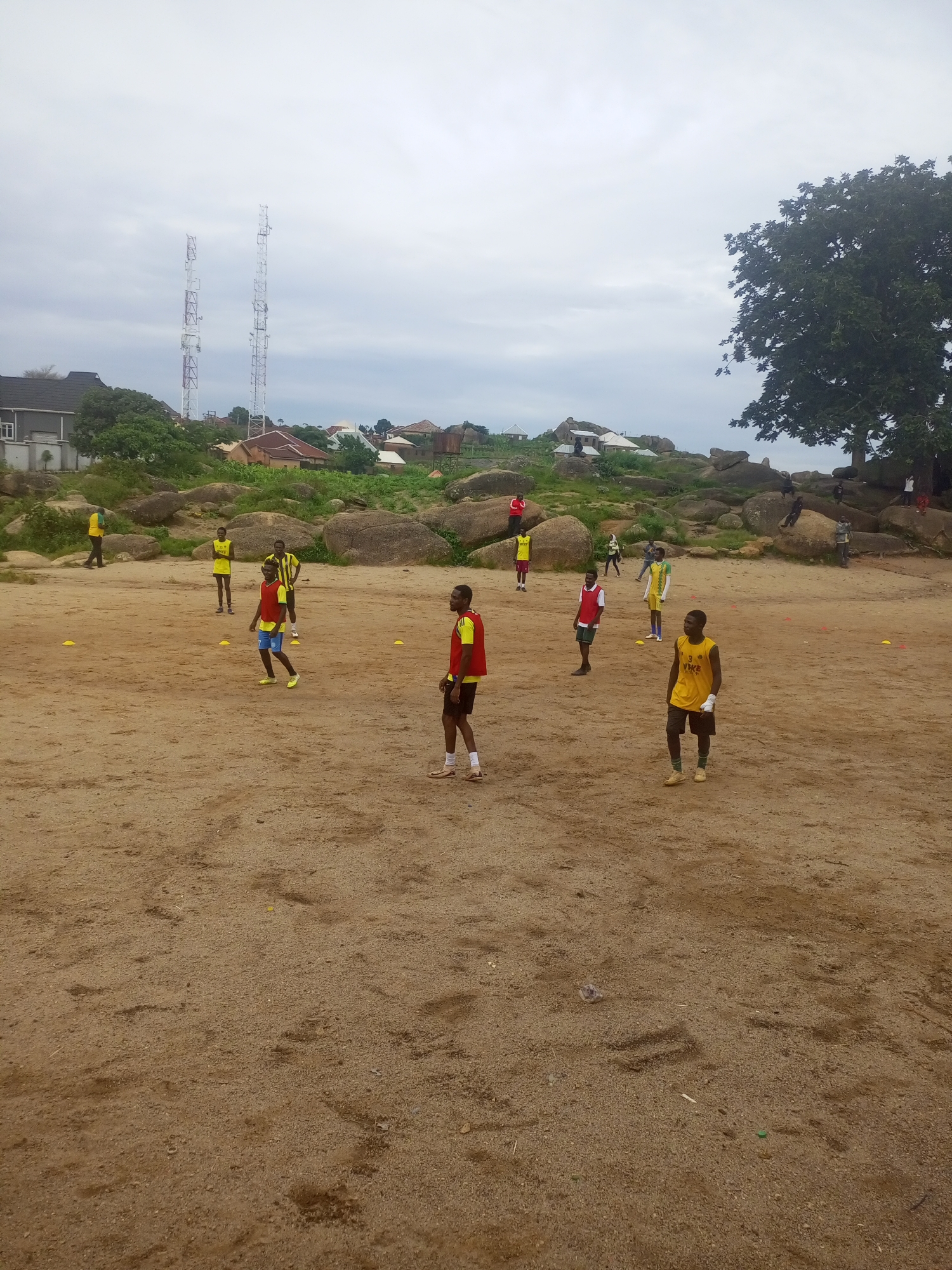 The image depicts an outdoor sports practice session, likely soccer training, taking place on a sandy ground. Several young men are present, dressed in athletic attire, some wearing yellow jerseys and shorts, others in red bibs over their clothes. They appear to be spread out across the field, engaged in drills or waiting their turn. In the background, a cluster of large rocks dominates the landscape, with a few scattered buildings and vegetation visible beyond them. Two prominent telecommunication towers stand tall against a cloudy sky, indicating the presence of infrastructure. The overall environment suggests a somewhat undeveloped or rural area on the outskirts of a city. Given the visual cues, particularly the rock formations and vegetation, this scene is consistent with locations found in and around the city of Jos, Nigeria.