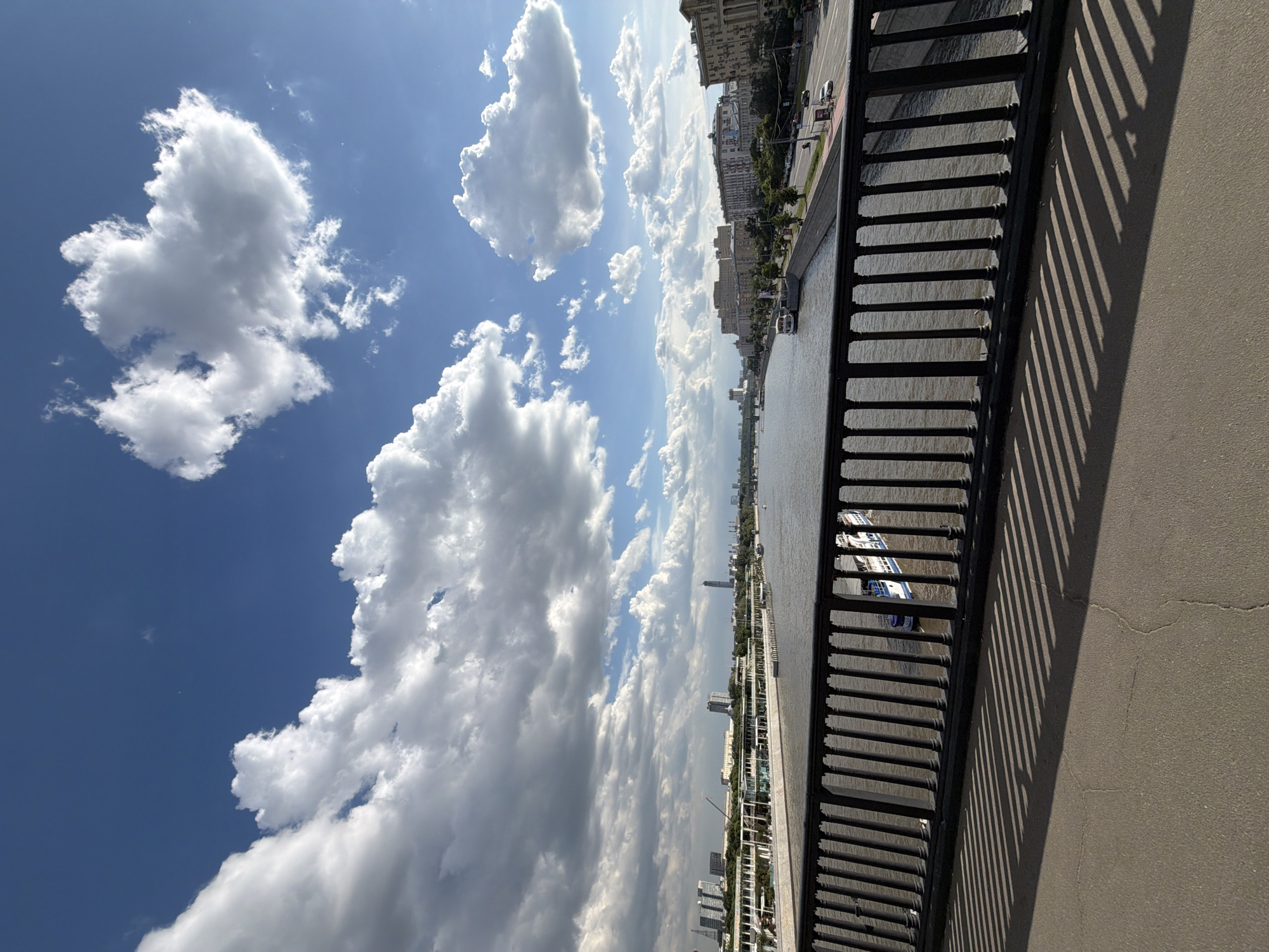 The image captures a scenic view of an urban riverfront under a bright, partly cloudy sky. The perspective is from an elevated vantage point, likely a bridge or an embankment walkway, looking across a river toward a city skyline.
The upper portion of the frame is dominated by a clear blue sky interspersed with large, bright white cumulus clouds, indicative of a sunny day. In the middle ground, a wide river or canal stretches horizontally, its surface reflecting the daylight. Along the opposite bank, a city panorama unfolds, featuring a dense arrangement of buildings. These structures vary in height and architectural style, including older, ornate multi-story buildings and more contemporary high-rises, all nestled alongside the river. Green foliage from trees lines parts of the embankment. Several boats, including one with distinctive white and blue elements, are visible on the water, while vehicles can be faintly seen on the road running parallel to the river.
In the immediate foreground on the right, a dark, ornate metal railing with vertical bars and posts casts long, diagonal shadows onto the light-colored concrete surface of the elevated platform. These sharp, well-defined shadows strongly suggest bright sunlight, indicating the time of day is likely mid-day to early afternoon. The overall atmosphere is one of a clear, pleasant day in a bustling city. Based on the provided context, the location is Moscow, Russia, likely along the Moscow River. No visible text is legible in the image.