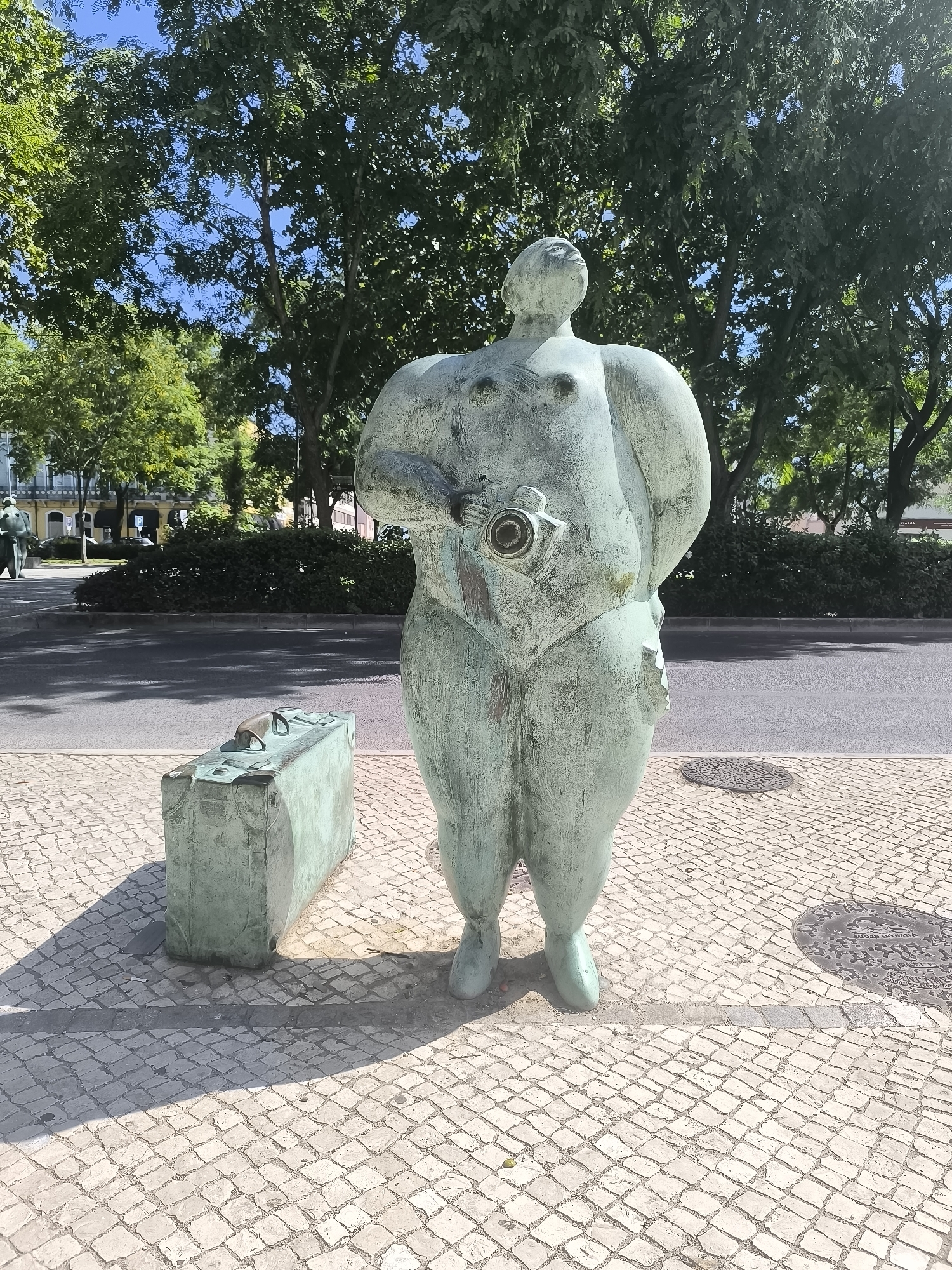 The image shows a bronze sculpture of a rotund figure holding a camera. The sculpture is situated on a cobblestone pavement next to a green suitcase. In the background, there is a street with a few cars and buildings. Lush green trees line the street, and the sky is a clear blue. The sculpture is likely located in Setúbal, Portugal, as indicated by the presence of Portuguese text on a manhole cover in the lower right portion of the image.