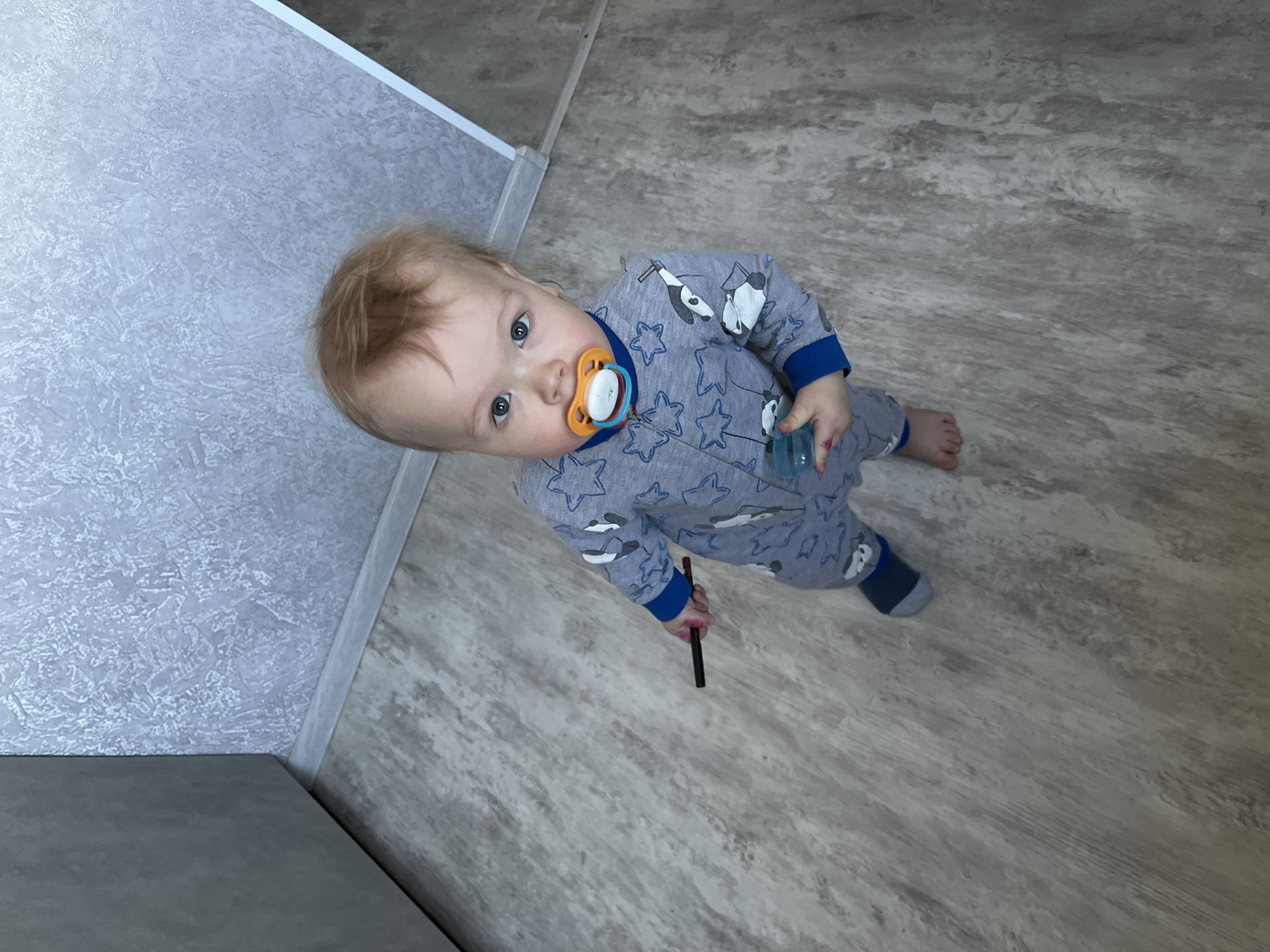 A young child, likely between one and two years old, stands on a linoleum floor. The child wears a gray pajama set with blue cuffs and a repeating pattern of stars and cartoon penguins. They have light blonde hair and blue eyes, and a pacifier in their mouth. The child holds a small, cylindrical object in one hand and a dark-colored pen or marker in the other. The floor is composed of gray and beige linoleum tiles. A section of gray wall or cabinet is visible in the upper left corner. The location of this photograph is unknown and there is no indication it was taken in Chaikovsky, Russia.