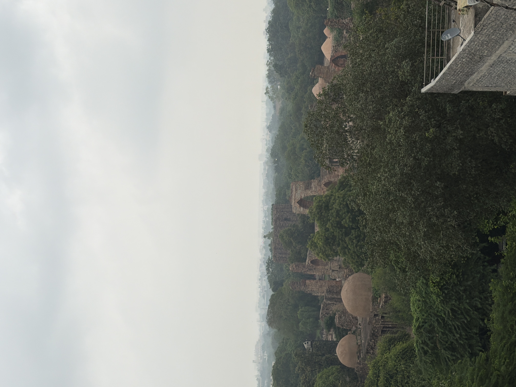 This is a high-angle, long shot of the Qutub Minar complex in Delhi, India, on an overcast day. The foreground features dense green foliage and a stone wall with a railing and a satellite dish. In the mid-ground, ancient red sandstone ruins, including arches and domed structures, are visible amidst more greenery. The background shows a hazy cityscape extending to the horizon. There are no people or animals visible in the image.