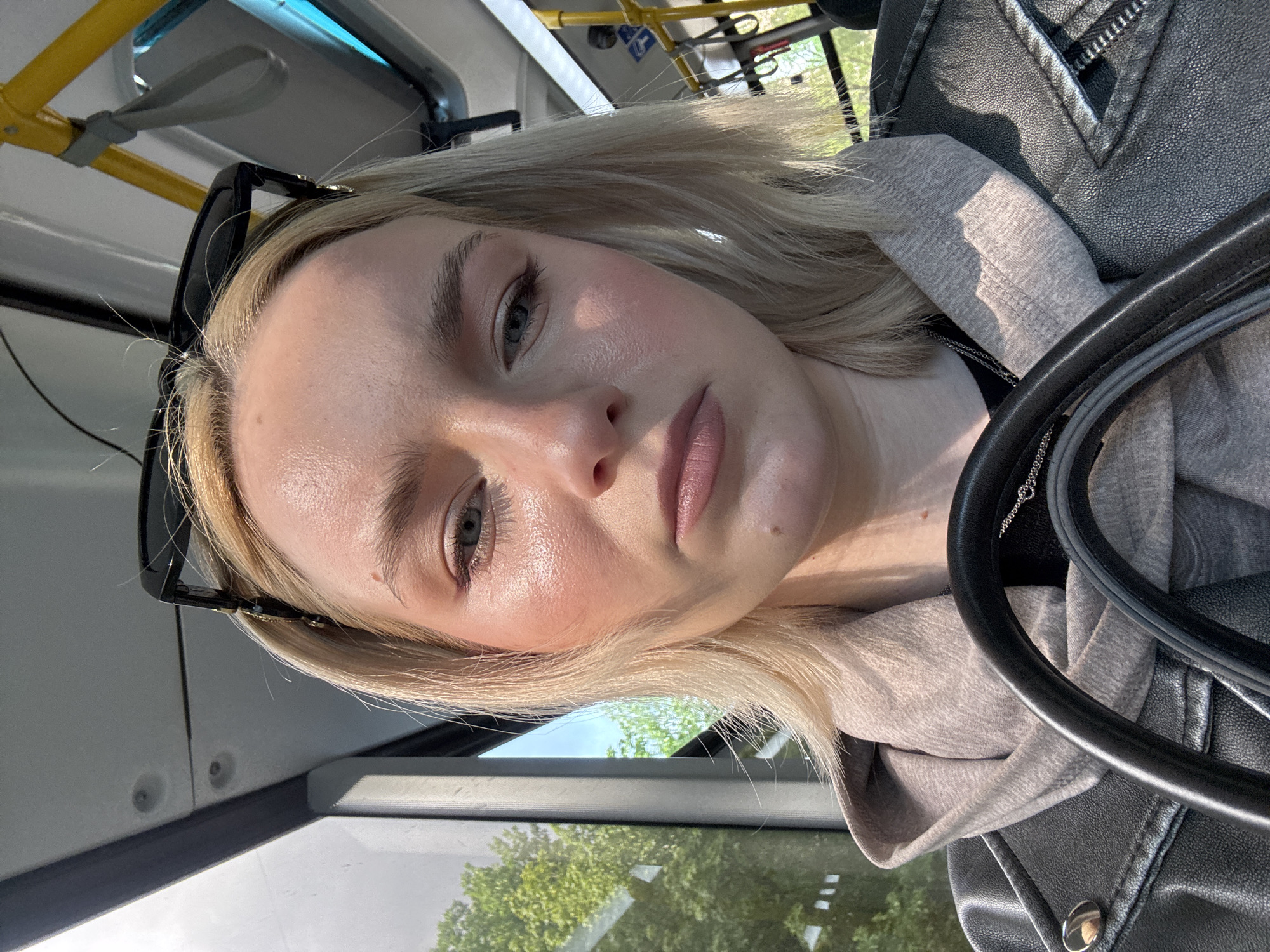 A young woman with blonde hair is taking a selfie on a bus. She is wearing a gray hooded sweatshirt and a black leather jacket. Sunglasses are perched on her head. The sun is shining, casting a warm glow on her face. Trees and a blue sky are visible through the bus window. The photo appears to be taken during the daytime. The scene is set inside a public bus, as indicated by the yellow grab handles and seating visible in the background. The woman's expression is neutral, and she seems to be looking directly at the camera. There is no visible text in the image.