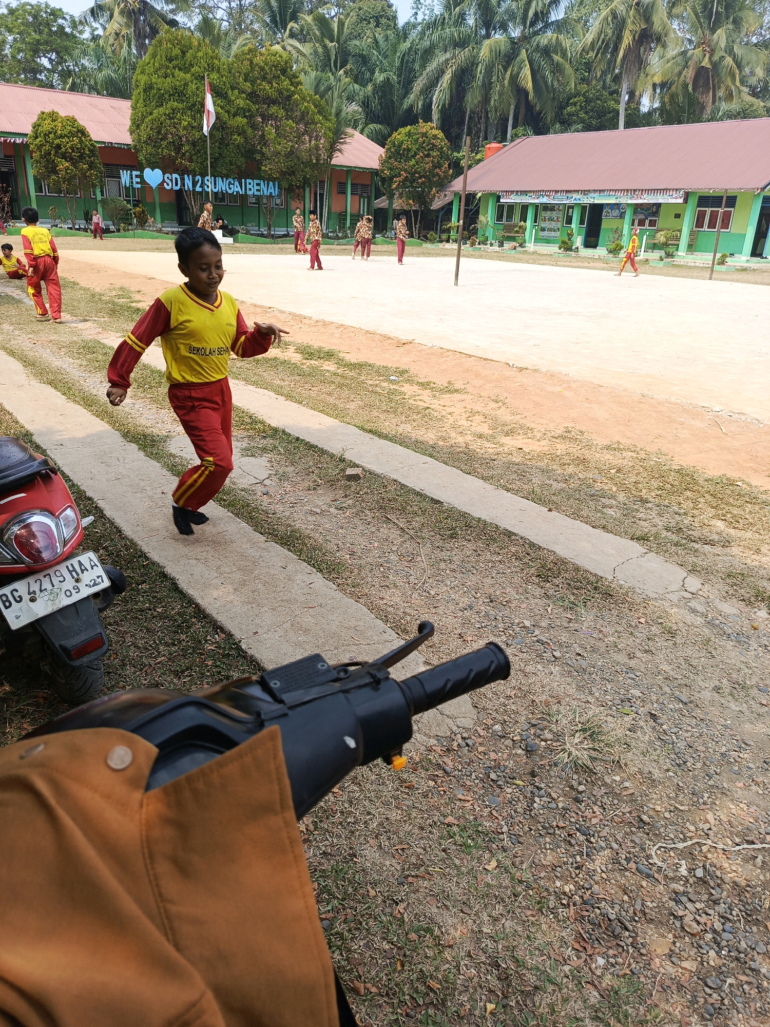 In the city of Megang Sakti IV, Indonesia, this image captures an outdoor scene at what appears to be a school. In the foreground, a portion of a brown jacket and handlebars of a motorcycle are visible. Behind this, a red motorcycle with a license plate reading "BG 4279 HAA 09-27" is parked on a concrete path. A young boy wearing a yellow and red uniform, with "SEKOLAH SEHA" visible on his shirt, is in motion, seemingly mid-stride or dance on the path.
The background features a school building with green walls and a reddish-brown roof, adorned with a sign that reads "WE ♡ SD N 2 SUNGAI BENAI". Several other children in similar uniforms are scattered across the sandy schoolyard, some engaged in activities. Palm trees and other lush vegetation surround the school, indicating a tropical or subtropical environment. The overall atmosphere suggests a bright, sunny day, likely during school hours or a school-related event.