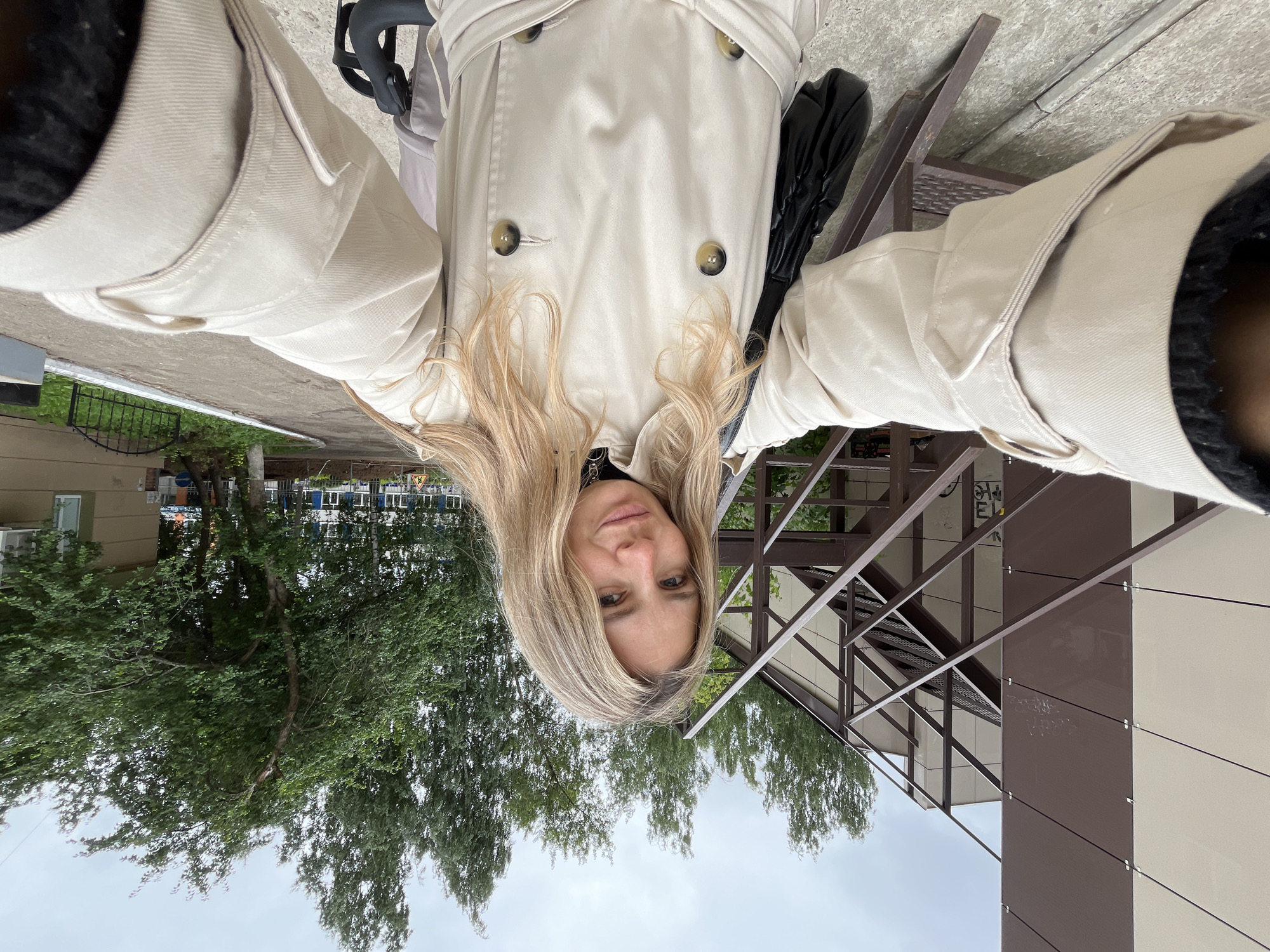 A selfie taken by a woman with blonde hair who is wearing a beige trench coat and a black bag. She is outside, with trees and a modern building in the background. The sky is overcast, suggesting it is daytime. The image is oriented vertically, with the woman's head at the bottom and her feet at the top. There is no visible text in the image.