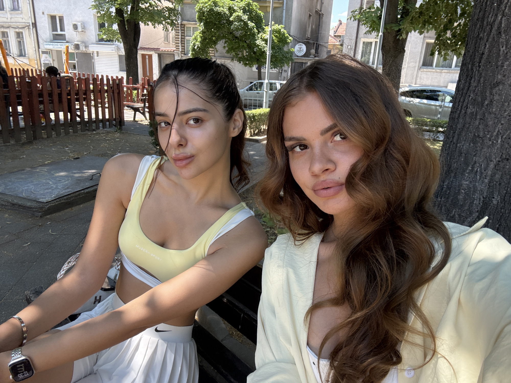 The media file shows a close-up, selfie-style shot of two young women sitting on a bench outdoors. The woman on the left has dark hair pulled back and is wearing a yellow sports bra and a white pleated skirt. She has a bracelet on her left wrist and a smartwatch. The woman on the right has long, wavy brown hair and is wearing a light-colored, possibly cream or pale yellow, button-up shirt over a white top. Both women are looking towards the camera with neutral expressions.
The background suggests an urban environment on a sunny day. Behind them, there are buildings, trees with green foliage, and a wooden fence enclosing a playground area where children can be seen. A silver car is parked in the background. The overall scene appears to be a public park or playground in a city.
There is some visible text on the yellow sports bra of the woman on the left, reading "MUSCULAR SPORT". The smartwatch on her wrist displays a digital time, though the numbers are difficult to read clearly. The weather appears to be warm and sunny, indicated by the bright lighting and clear sky. The location context provided is Plovdiv, Bulgaria.