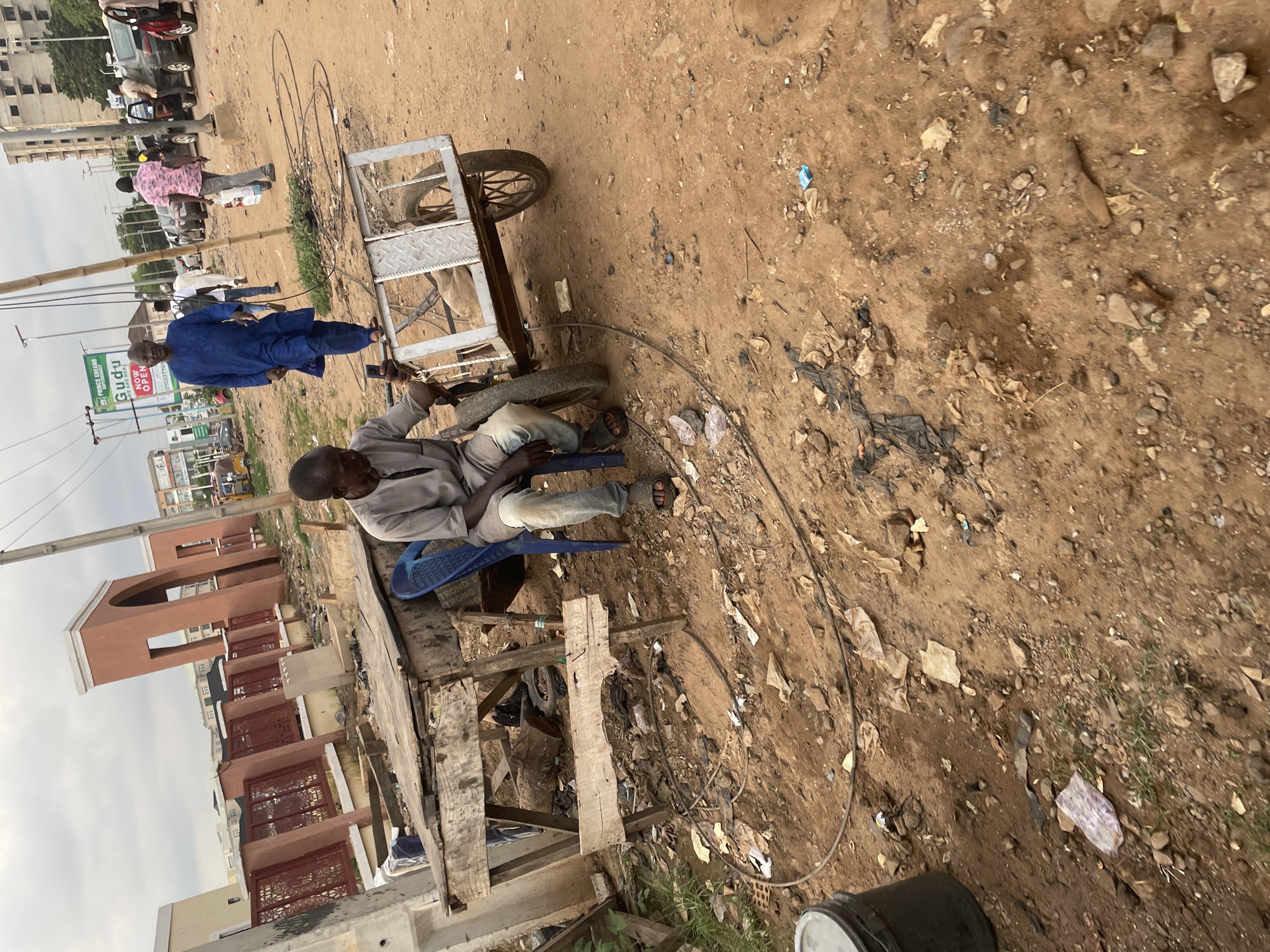 The image depicts a street scene in Abuja, Nigeria. The main subjects are two men and a cart. One man, wearing a blue embroidered garment, is standing near the cart. He appears to be observing the other man. The second man, dressed in a grey shirt and pants, is seated on a blue plastic chair beside the cart. He seems to be engaged in some activity with the cart, possibly working on it or preparing to move it. The cart itself is a simple, two-wheeled metal structure with a wooden platform, possibly used for transporting goods.
The setting is an unpaved, dusty area that looks like a roadside market or a construction site. The ground is covered with dirt, rocks, and debris, including plastic bags and other discarded items, suggesting a somewhat informal and possibly less affluent area. In the background, there are buildings under construction, with exposed brickwork and scaffolding visible, indicating ongoing development. There are also a few vehicles parked on the side of the road and some people in the distance, adding to the sense of a busy, active environment.
The time of day appears to be daytime, with overcast skies suggesting cloudy weather. The overall mood is neutral, focusing on the everyday activities of the people in the scene. There is a visible sign with text that reads "GUDU PUBLISHING" and "NOW OPEN" with a phone number. The scene is characteristic of many urban areas in Nigeria, where street vendors and informal labor are common.