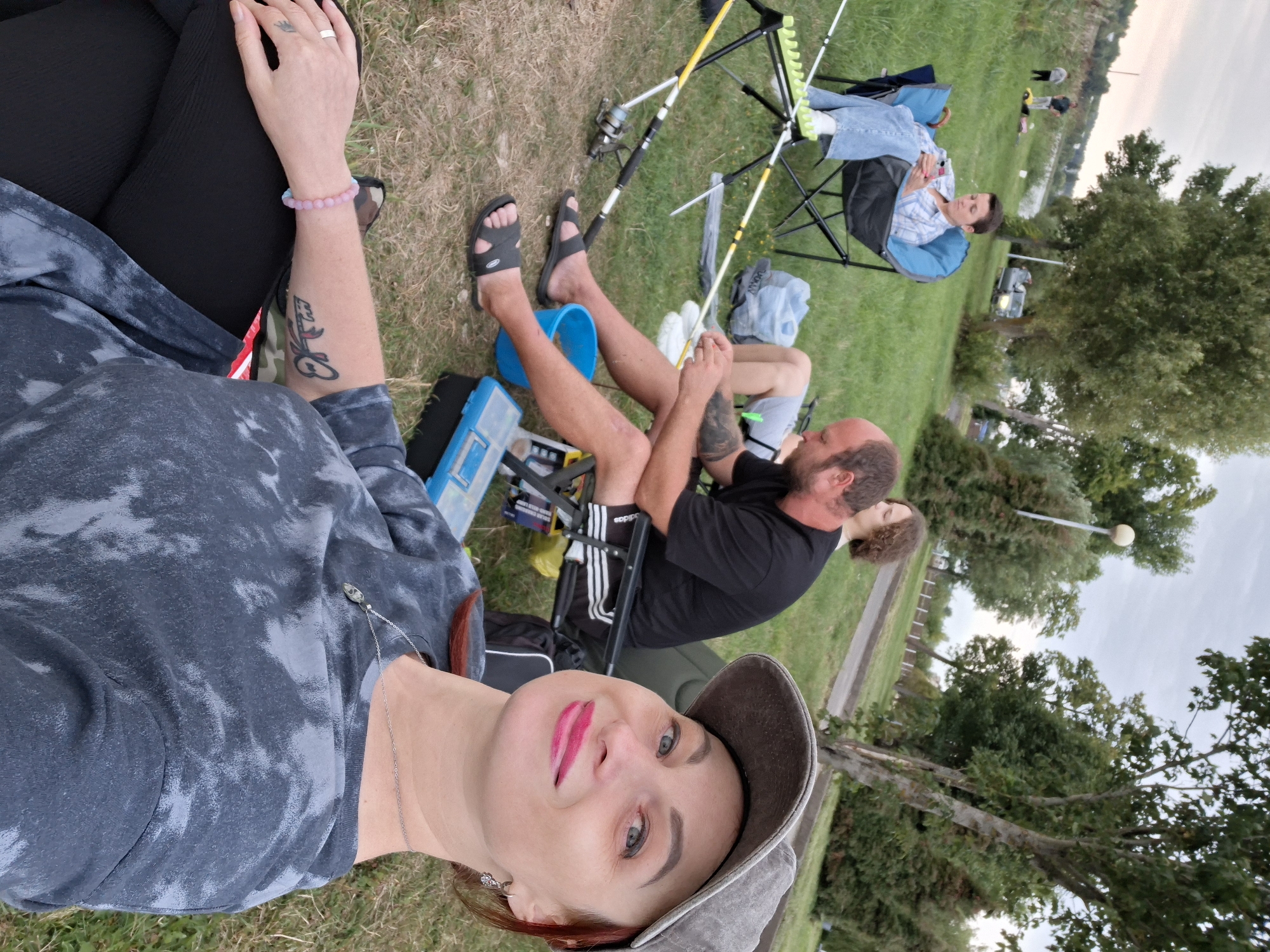 In Myadzyel, Belarus, on a grassy area near a body of water, a group of people are engaged in fishing. A woman in the foreground, wearing a gray tie-dye t-shirt, a gray baseball cap, and lipstick, is taking a selfie. She has a bracelet on her left wrist and a ring on her ring finger. Behind her, a man with a beard, wearing a black t-shirt and shorts, is sitting in a camping chair with fishing rods in front of him. To his left, a woman in a blue plaid shirt and jeans is seated in a similar chair, looking at her phone. Another person is visible in the background. The environment includes trees, grass, a fishing rod holder, buckets, and a fishing tackle box. The sky is overcast.