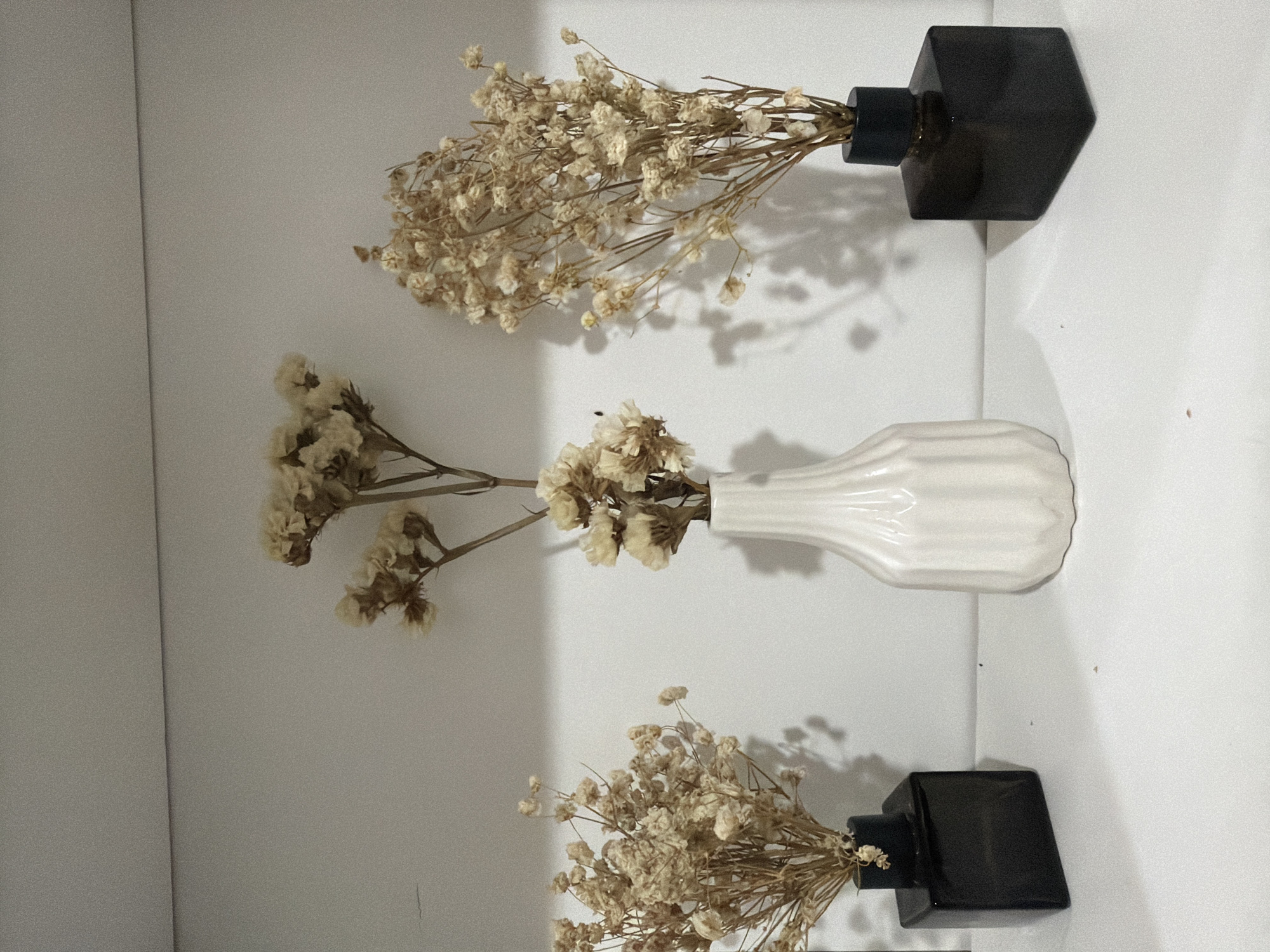 The image shows three vases with dried flowers on a white surface. Two vases are dark, possibly black or dark brown, with a geometric, somewhat hexagonal shape. The third vase is white with vertical ridges. The dried flowers are light in color, appearing beige or cream. The scene is indoors. The angle and lighting suggest it's a shelf or display area. No people are visible. There is no specific information in the image to suggest the location is Davao City, Philippines.
