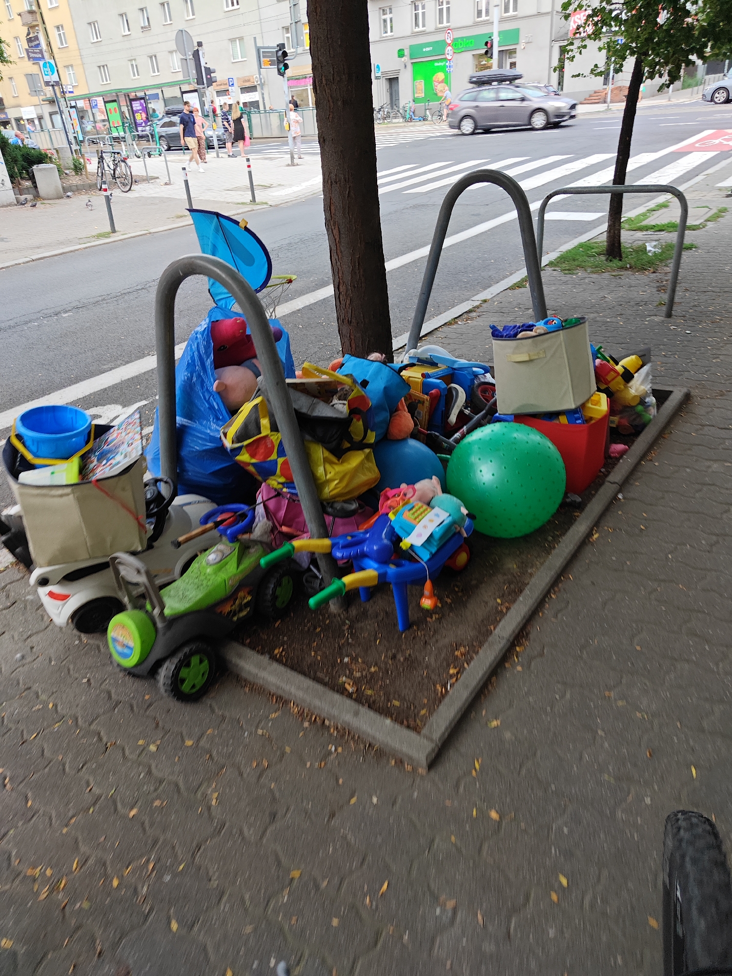 This image depicts a street scene in Poznan, Poland, specifically a sidewalk area near a road with a crosswalk. The foreground is dominated by a collection of children's toys that appear to have been left out, possibly for collection or disposal. These include a large blue plastic bag filled with stuffed animals, a green ride-on toy car, a large green inflatable ball, a red storage bin, and a beige fabric storage cube containing more toys. A blue plastic bucket and a disassembled blue umbrella are also visible. In the background, a road with a crosswalk is visible, with a few cars and pedestrians present. Buildings line the street, suggesting an urban environment. A tree trunk is prominent in the right half of the image, and a bicycle rack is present. The overall impression is of an outdoor urban setting with a somewhat untidy arrangement of discarded children's items.