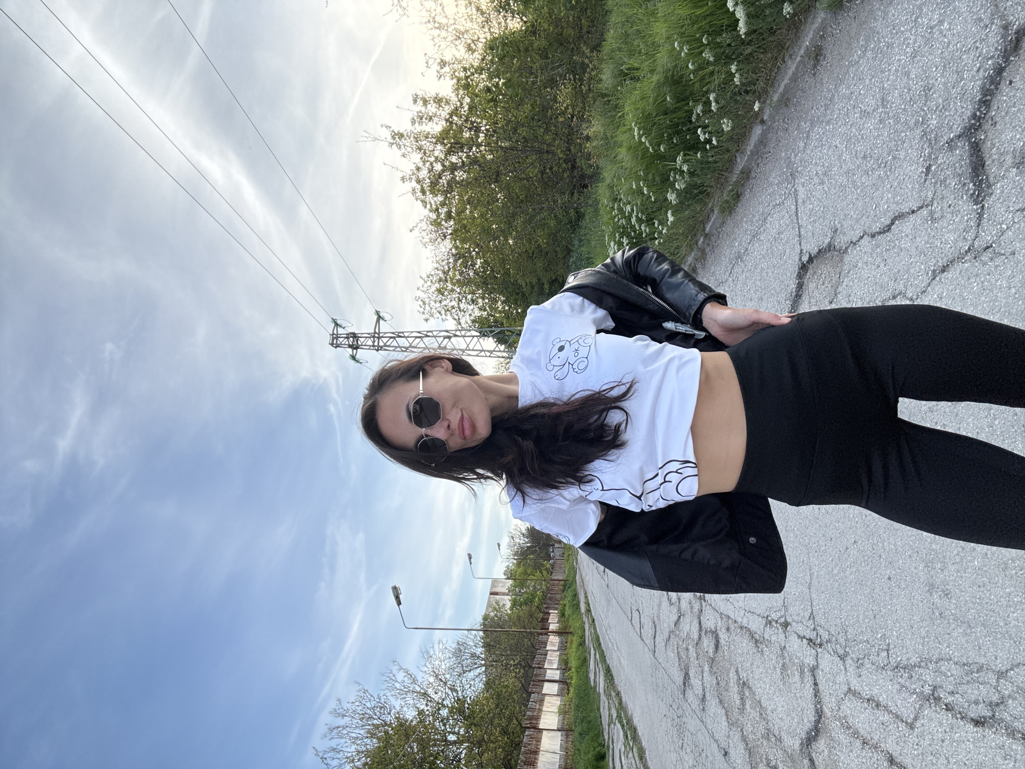 A young woman stands on a cracked asphalt road in what appears to be an urban fringe or industrial area in Pleven, Bulgaria. The image, which is rotated, depicts her in a three-quarter pose, looking directly at the camera with a neutral expression, possibly a slight smirk, from behind dark sunglasses. She has long, dark hair flowing over her shoulders.
She is dressed in a white crop top featuring a black cartoon-style dog graphic with "X O" near it, black leggings, and a black leather jacket casually draped over her shoulders. Her left hand is placed on her hip.
The setting includes a paved path or road extending into the distance, flanked by green grass and small white wildflowers on one side, and a metal fence line with indistinct buildings or structures further along on the other. Two streetlights are visible down the road. Overhead, power lines crisscross the bright sky, connecting to a metal pylon structure visible on the left side of the frame. The sky is largely clear with scattered wispy clouds, suggesting late afternoon or early evening lighting.
