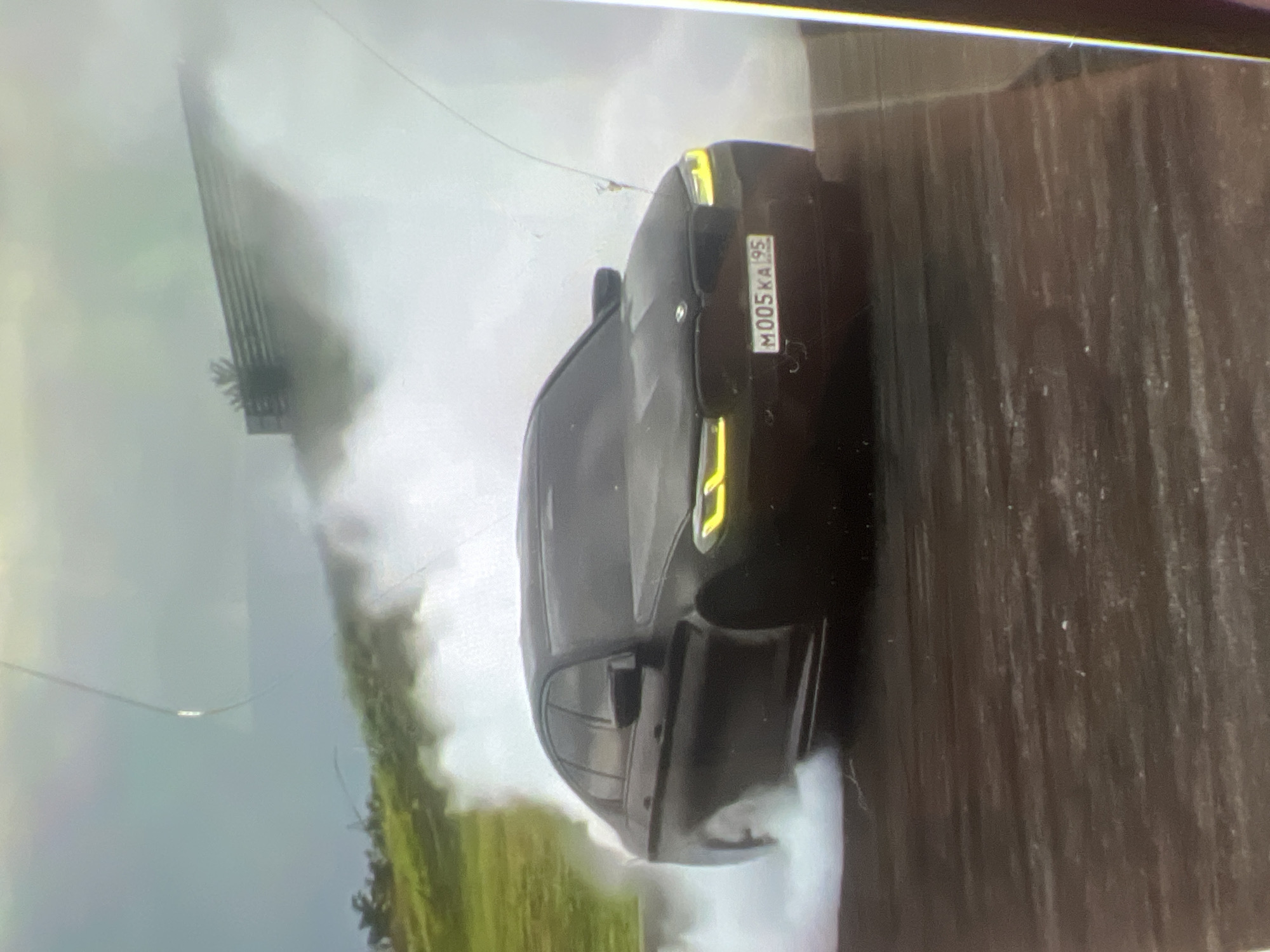 A black BMW sedan is performing a drift in what appears to be an outdoor, possibly urban or track environment. The car is emitting a large amount of white smoke from its tires, indicating aggressive maneuvering. The license plate of the car is visible and reads "M005KA95". The background shows a grassy incline with some trees and a fence-like structure partially obscured by smoke. The ground beneath the car appears to be paved or a similar dark surface. The sky is overcast. No people are clearly visible in the image. The overall scene suggests a dynamic and possibly professional driving event. This image is not specific to the city of Kara-Dıykan, Kyrgyzstan.