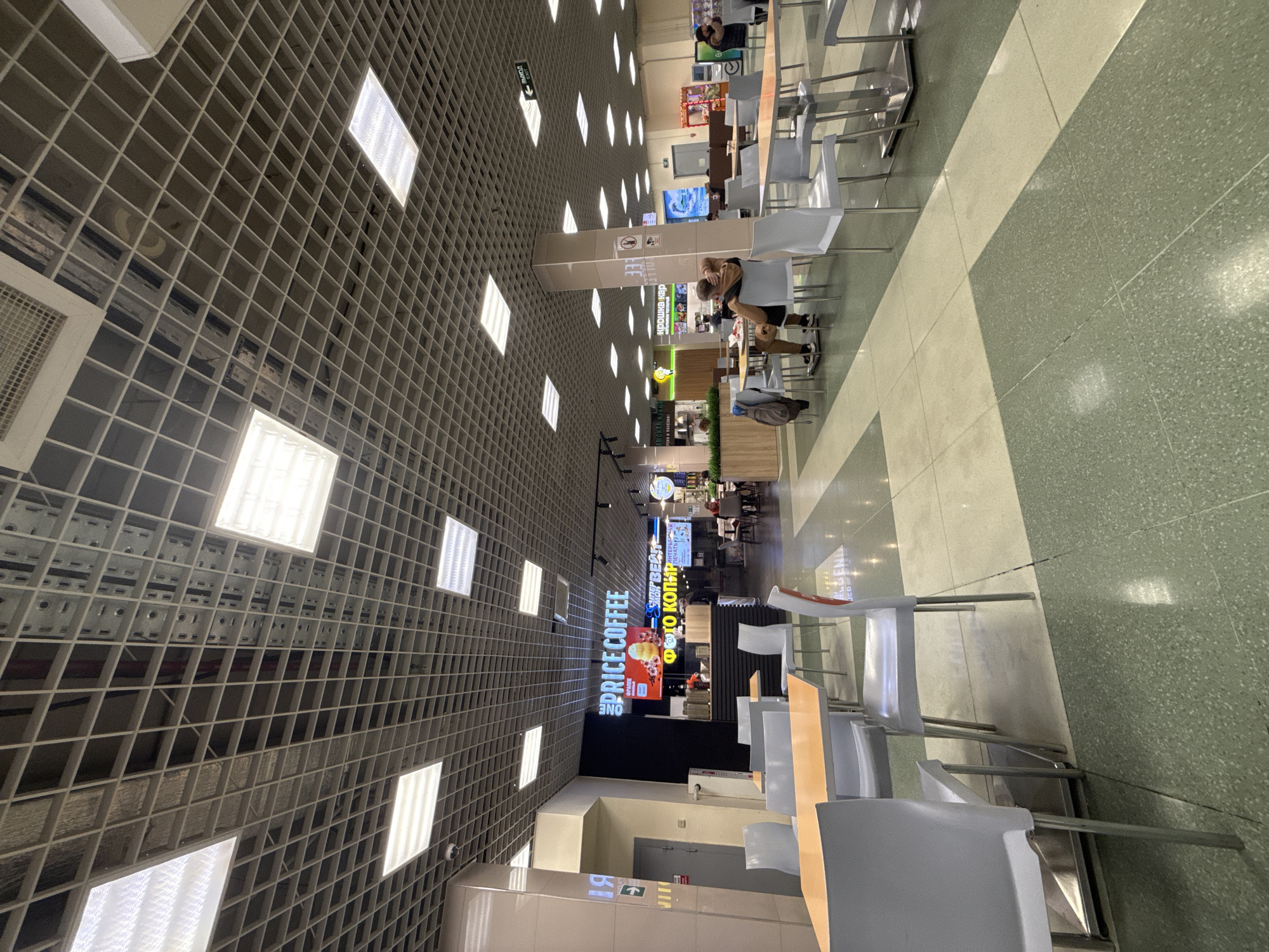 The image shows an interior view of a food court in what appears to be a shopping mall in Mytishchi, Russia.
**Environment:** The floor is a combination of light and dark green tiles. The ceiling is a grid-like structure with fluorescent lights evenly spaced. Various food vendors are visible in the background.
**Objects:** Tables and chairs are arranged in the foreground. Signs for businesses are visible.
**People:** At least one person is seen sitting at a table.
**Location:** Based on the signage and details, this location is in Mytishchi, Russia.