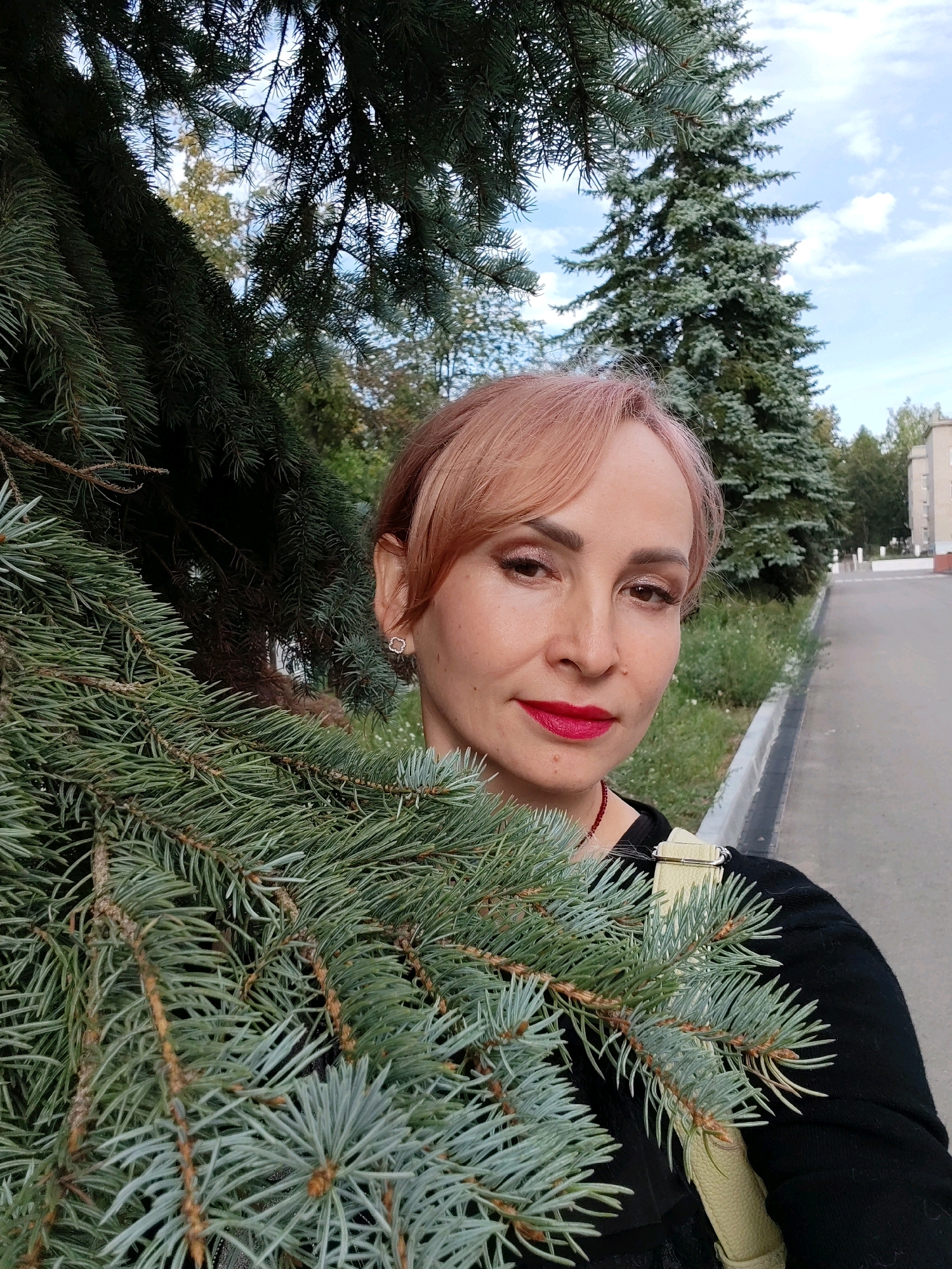 An outdoor, medium shot selfie shows a light-skinned woman with short, light pink hair and bright red lipstick. She is positioned in front of a blurry background of evergreen trees, a paved walkway with a curb, and a light-colored building. A large branch of a pine tree with blue-green needles is in the foreground, obscuring part of the woman's face and body. The woman is wearing a black sweater and a light yellow purse strap is visible over her right shoulder. She appears to be outdoors in a park or residential area, potentially in the city of Chaikovsky, Russia, given the presence of mature coniferous trees and a building that resembles typical Soviet-era architecture. The sky is partly cloudy.