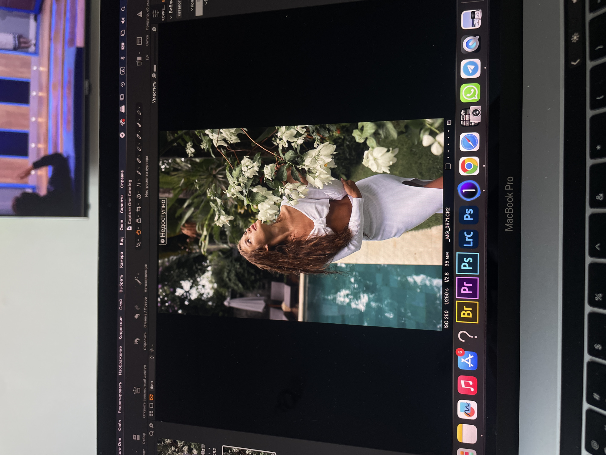 The image is a photograph of a MacBook Pro screen. The screen displays the Capture One photo editing software with a vertical portrait of a woman in a white dress next to a swimming pool and flowering plants. The camera settings, ISO 250, 1/250 s, f/2.8, and 35mm lens are displayed. There are application icons visible in the dock. A portion of a television screen is visible to the left of the laptop, showing a stage with two people on it. There is no information available to determine if this image was taken in the city of Kazan, Russia.