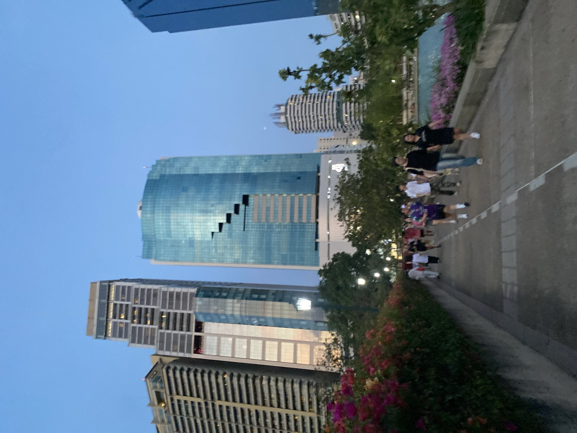The image shows a street scene in Bangkok, Thailand at dusk. Several skyscrapers are visible, with one prominent building featuring a distinct cutout design on its facade. A group of about ten people are walking along a paved pathway beside a flower bed with pink and red blossoms. Street lamps line the path, and trees are interspersed among the buildings. The sky is a clear blue.