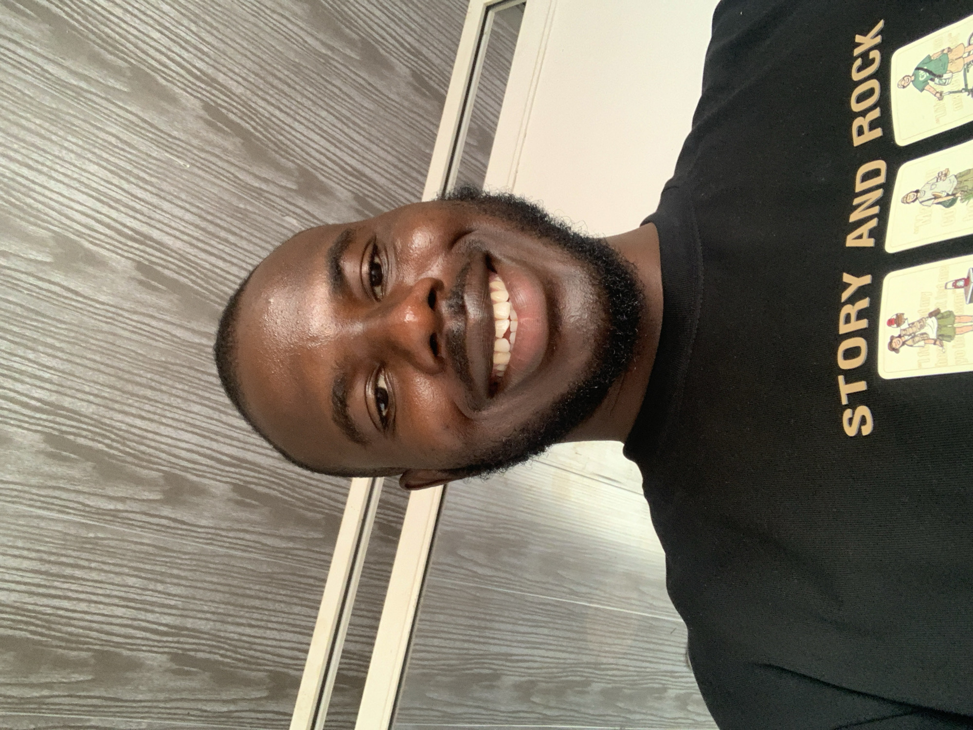 A smiling dark-skinned man with a close-cropped haircut and a beard is wearing a black T-shirt with the words "STORY AND ROCK" and images on it. The background consists of a grey-toned wall with horizontal textured lines, and a white door frame. The location and city details are not apparent in this image.