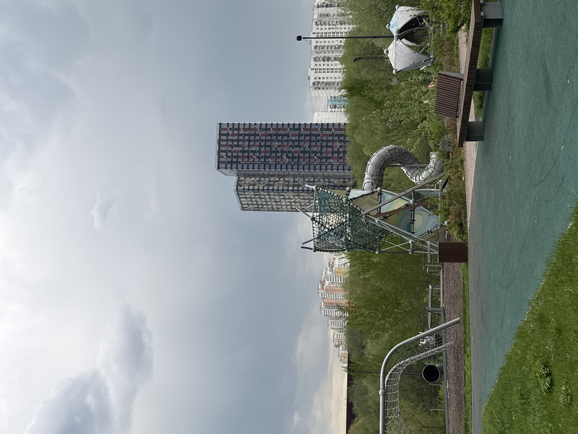 The image captures a view of a playground adjacent to residential high-rise buildings, situated within the city of Moscow, Russia. The foreground features typical playground equipment including a climbing structure with a net, a winding tube slide, and a soccer goal. A bench is placed alongside the playground area. The background is dominated by rows of tall apartment buildings, characteristic of urban Moscow. The sky is overcast, giving the scene a muted, somewhat gray tone. The overall environment suggests a park or recreational area integrated within a residential neighborhood. No people are visible in the frame.
