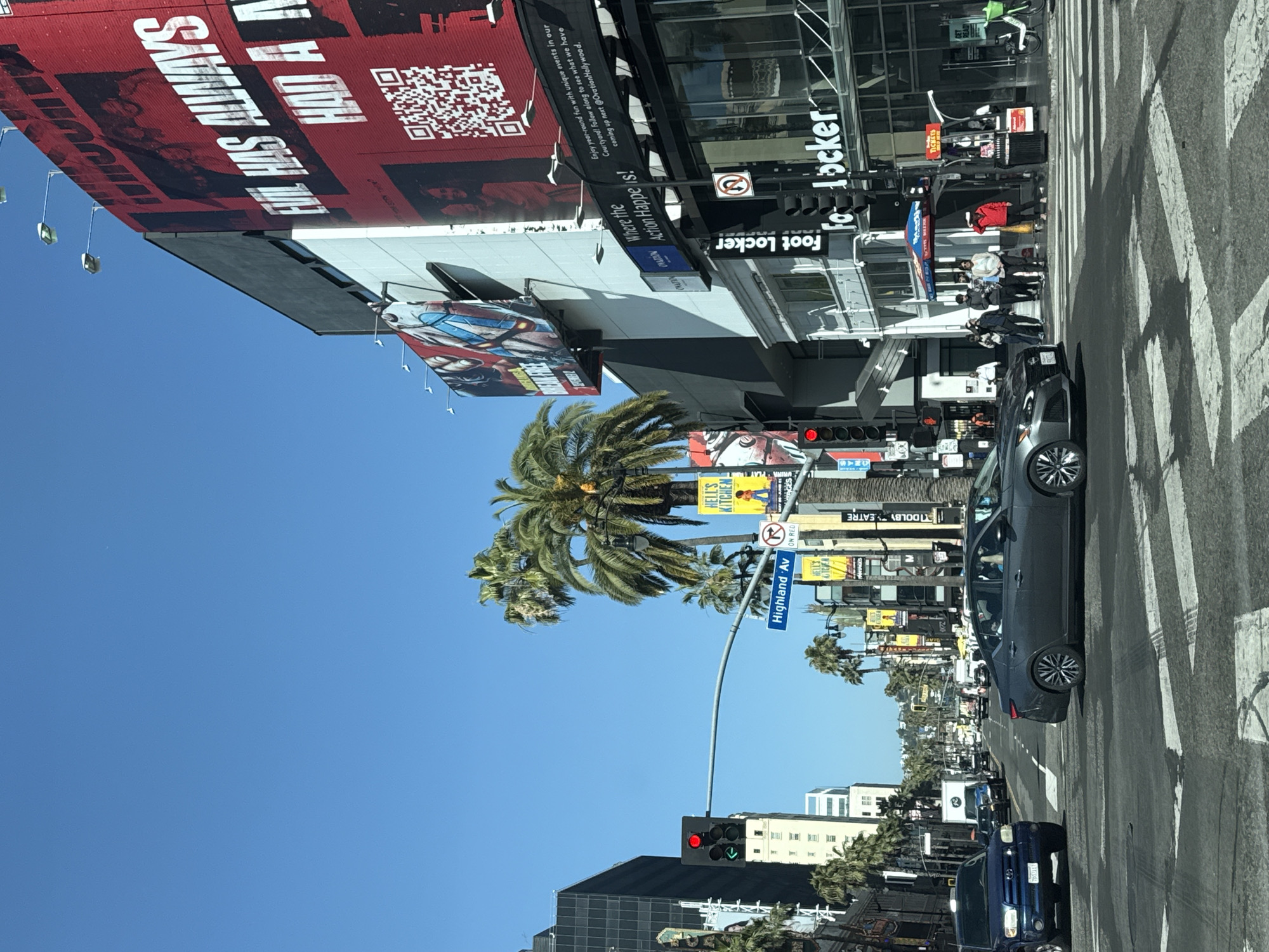 The image captures a vibrant daytime street scene in Hollywood, Los Angeles, under a clear blue sky, suggesting bright, sunny weather. The perspective is looking down a busy street, likely Highland Avenue, indicated by a prominent blue street sign.
Commercial buildings line both sides of the street. On the left, a large red billboard features bold white text, partially reading "ALWAYS HOLY A VA" and displaying a QR code. Below it, a banner invites engagement with "Ovation Hollywood," stating, "Enjoy year-round fun with unique events in our Courtyard! Follow along to see what we have coming up next @Ovation Hollywood." and "Where the Action Happens!". The "Foot Locker" brand is conspicuously displayed on multiple signs on the adjacent building. Other visible businesses or signs include "HELL'S KITCHEN," a partially visible "Dolby Theatre," and "PLAY ON RED."
The street is active with vehicles; a dark grey sedan is prominent in the foreground, and a blue pickup truck is further down the road, with more traffic in the distance. Pedestrians are visible near a crosswalk, with a group gathered on the sidewalk in front of the Foot Locker store. A traffic light shows a red signal for straight-ahead traffic and a green arrow, indicating that turns are permitted.
Numerous palm trees, characteristic of Southern California, are interspersed throughout the urban landscape. The scene depicts a typical moment of bustling urban life in a well-known commercial and entertainment district.