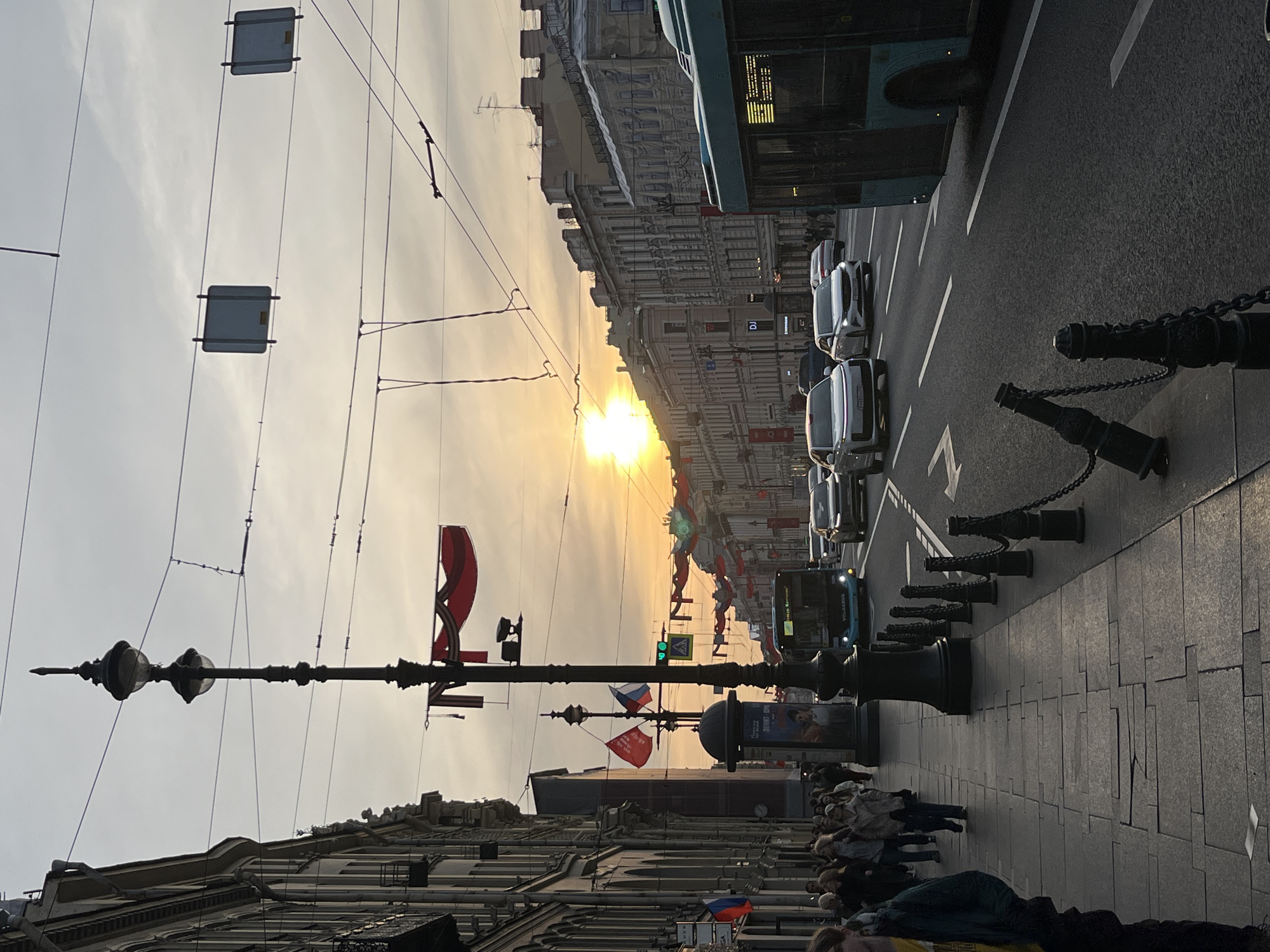 The image captures a street scene in St. Petersburg, Russia, during what appears to be a public celebration or event. A crowd of people is gathered on the sidewalk along the street. The road is filled with cars and buses. Red flags are prominently displayed along the street, including those of Russia. Overhead wires span the street, and a large lamppost stands in the foreground. The image is taken at dusk, with the setting sun visible in the distance. The buildings along the street are classic European style, with ornate facades and many windows.