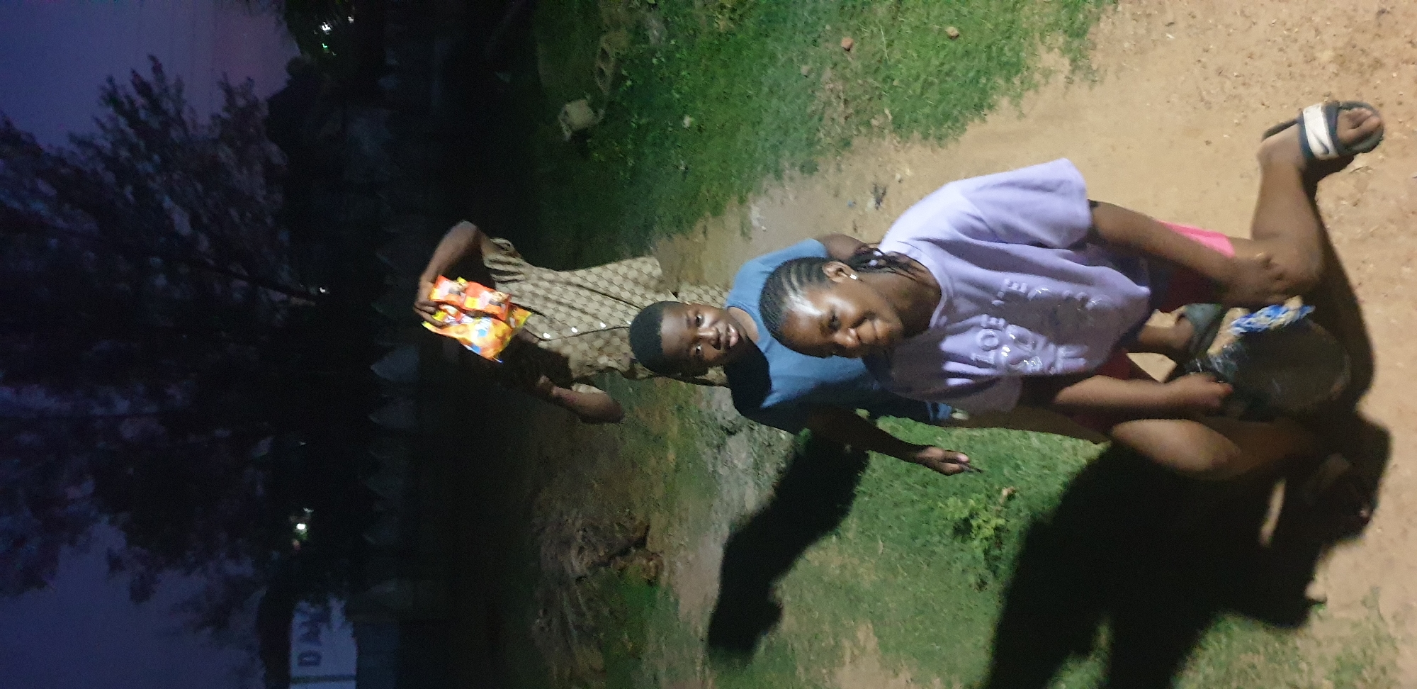 This image captures three individuals outdoors at dusk or nighttime in the city of Barden Yana, Nigeria. The scene is illuminated by an unseen artificial light source, casting long shadows. In the foreground, a young female with cornrows is kneeling on a reddish-brown dirt path, wearing a light purple t-shirt with "LOVE" printed on it, pink shorts, and a dark slide sandal on her left foot. She is holding a dark, possibly plastic, bucket or container adorned with a blue and white patterned cloth. Her expression is a slight smile directed towards the viewer. Directly behind her, a young male, appearing to be pre-teen, stands on the same dirt path, wearing a blue t-shirt and looking forward with a neutral expression. Further back on the path, another male figure stands, wearing a light brown patterned short-sleeved shirt. This individual is holding a translucent plastic bag filled with colorful snack packages above his head, obscuring his face. The ground consists of a dirt path bordered by green grass. In the distant background, dark tree silhouettes are visible against a dim twilight sky, along with faint distant lights. A partially visible white sign with "DA" is present on the far left. A row of small, light-colored, pointed structures or markers is discernible in the deeper background.