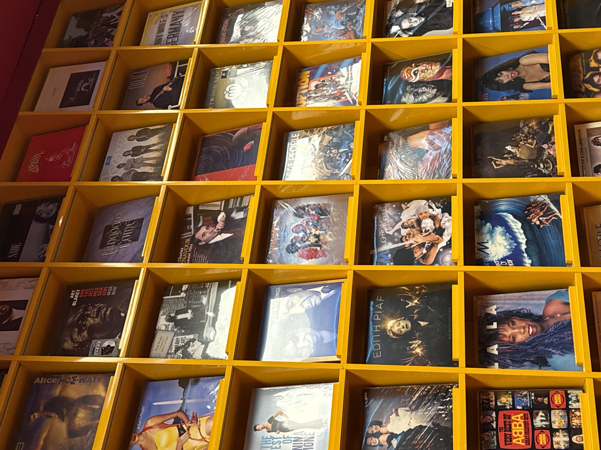 This image depicts a wall of vinyl records displayed on yellow shelves. The records are organized in a grid pattern, with each shelf holding multiple records. The records feature various artists and album covers, suggesting a music store or a private collection. The lighting appears to be artificial, casting shadows on some of the records. The background is a plain wall. The image does not contain any information that would suggest a specific location in Krasnodar, Russia.