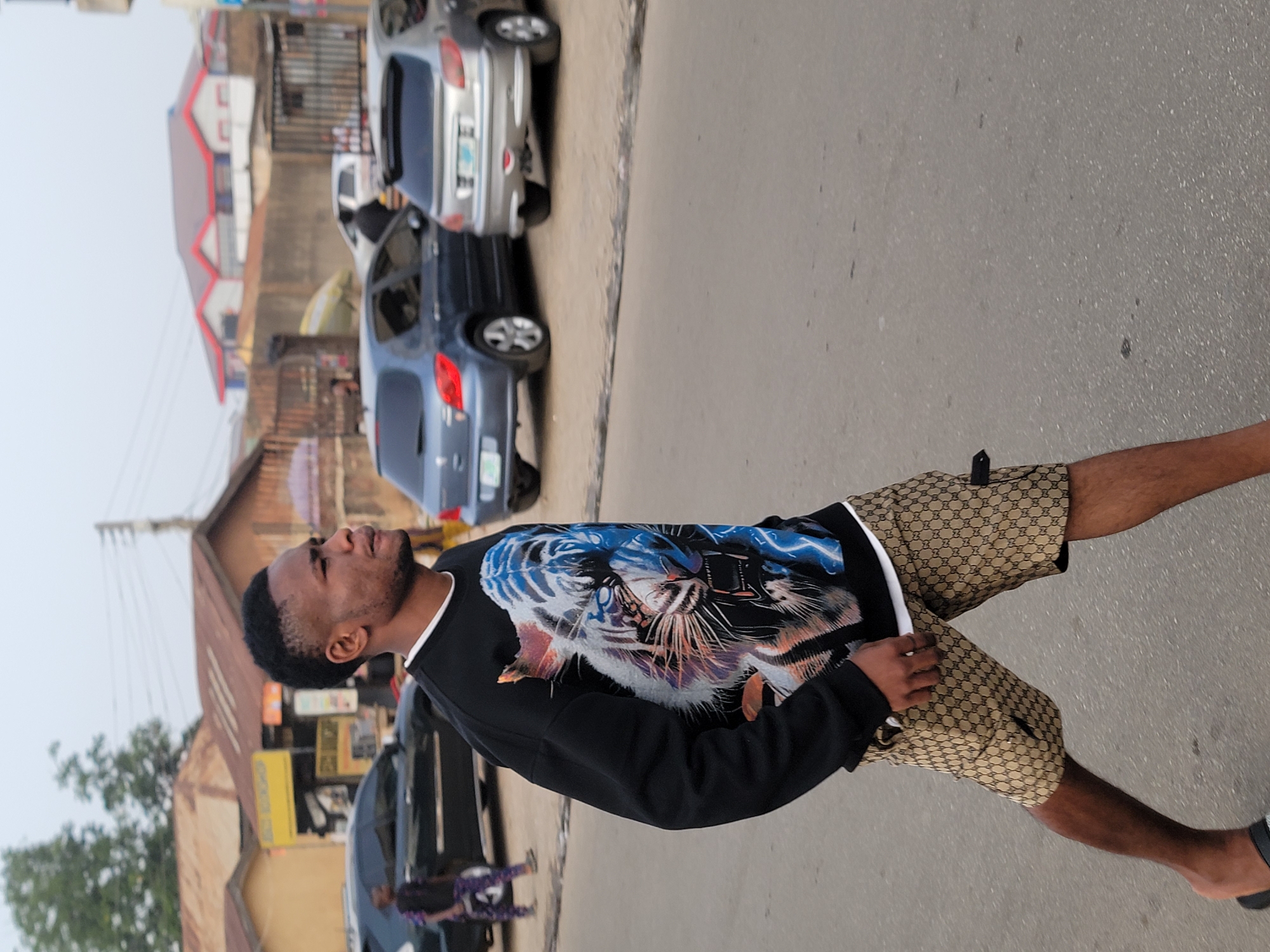 A man is standing on a street in what appears to be a city. He is wearing a black long-sleeved shirt with a tiger design and designer shorts. He appears to be looking up. Several cars are parked along the side of the street, and there are buildings visible in the background. Based on other data points available, this is assumed to be in Jos, Nigeria.
