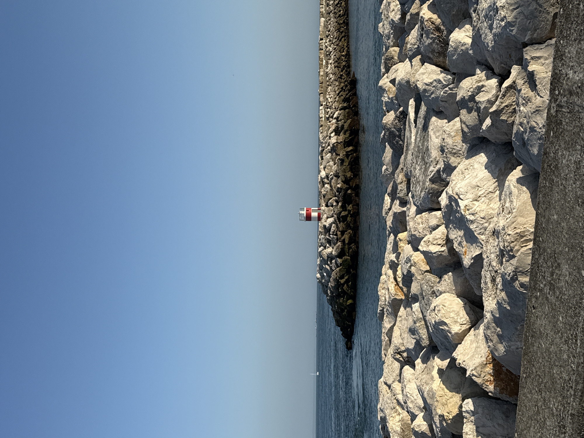 This is a vertical, eye-level shot of a breakwater made of large rocks, with a small lighthouse at the end, on a clear, sunny day. The lighthouse is red and white and stands on a rocky outcrop. The sea is calm and blue, with a few sailboats in the distance. The foreground is filled with large, light-colored rocks. The scene is located at the marina of Oeiras, Portugal.