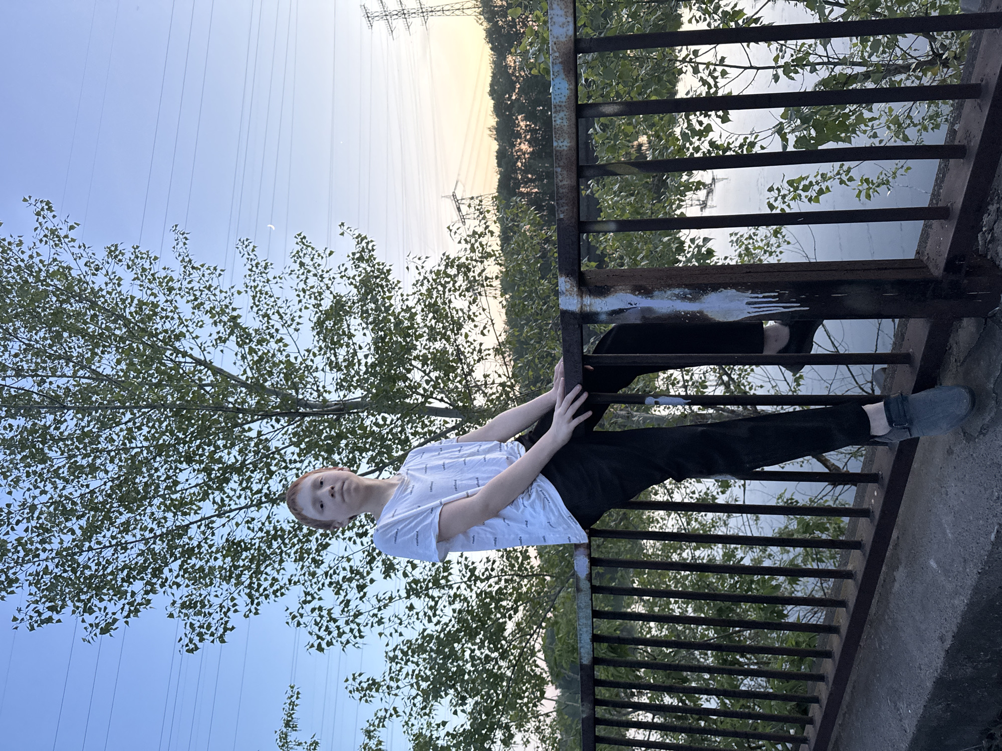 A young person is balancing on the rusted railing of a bridge or overpass. The person is light-skinned with red hair, wearing a white t-shirt with words printed on it and black pants. They are holding onto the railing with both hands. Below the railing, trees and what appears to be water are visible. Overhead power lines stretch across the sky. The location is possibly in or near Chaikovsky, Russia. The time of day is likely late afternoon or early evening due to the lighting.