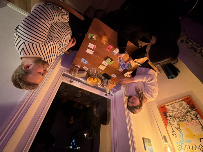 An interior photograph, captured from an elevated perspective, displays three individuals gathered around a light brown wooden table, engaged in a card game. The setting is an indoor space, characteristic of a private residence, at night.
On the left, a subject with dark hair, seen from the back, holds multiple playing cards. To their right, a female subject with light brown hair, wearing a light-colored textured sweater, holds or places cards on the table. On the right, a male subject with light brown hair and a beard, dressed in a white and dark striped shirt, looks to his left, away from the immediate game activity.
The table features numerous colorful playing cards, some face-up, indicating an ongoing game. Also present are two drinking glasses (one containing an amber liquid with ice), a small white bowl filled with yellow citrus fruits, a clear bottle, and a small white electronic device.
The room is illuminated by warm artificial light, with a prominent bare incandescent bulb visible on the upper left wall. A large poster, depicting an urban scene with "Louis Vuitton Home Printemps Ete 2021" text, hangs on the left wall above a dark rolled-up mat. A smaller framed picture is mounted higher on the same wall. A large window with white framing occupies the right wall, reflecting the dark exterior night sky and an internal blue, monitor-like light source, along with a faint human silhouette.
No specific visual identifiers within the image directly confirm its location as London, United Kingdom. However, the scene is consistent with an urban dwelling.