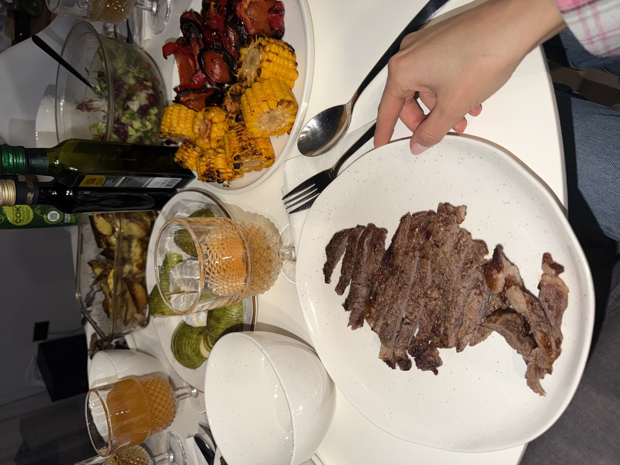 A top-down view shows a dining table with various food items and utensils. A white plate in the lower right corner holds sliced beef. A fork and spoon rest on the table near the plate. Another plate at the top of the image contains grilled corn on the cob and red bell peppers. A salad is visible in a bowl in the upper left corner, next to bottles of oil. Other items on the table include a glass of light-colored liquid, a bowl, and what appear to be other dishes or food containers. A person's hand reaches towards the plate with the beef. The context suggests a meal setting. There is no indication of the location being Krasnoyarsk, Russia.