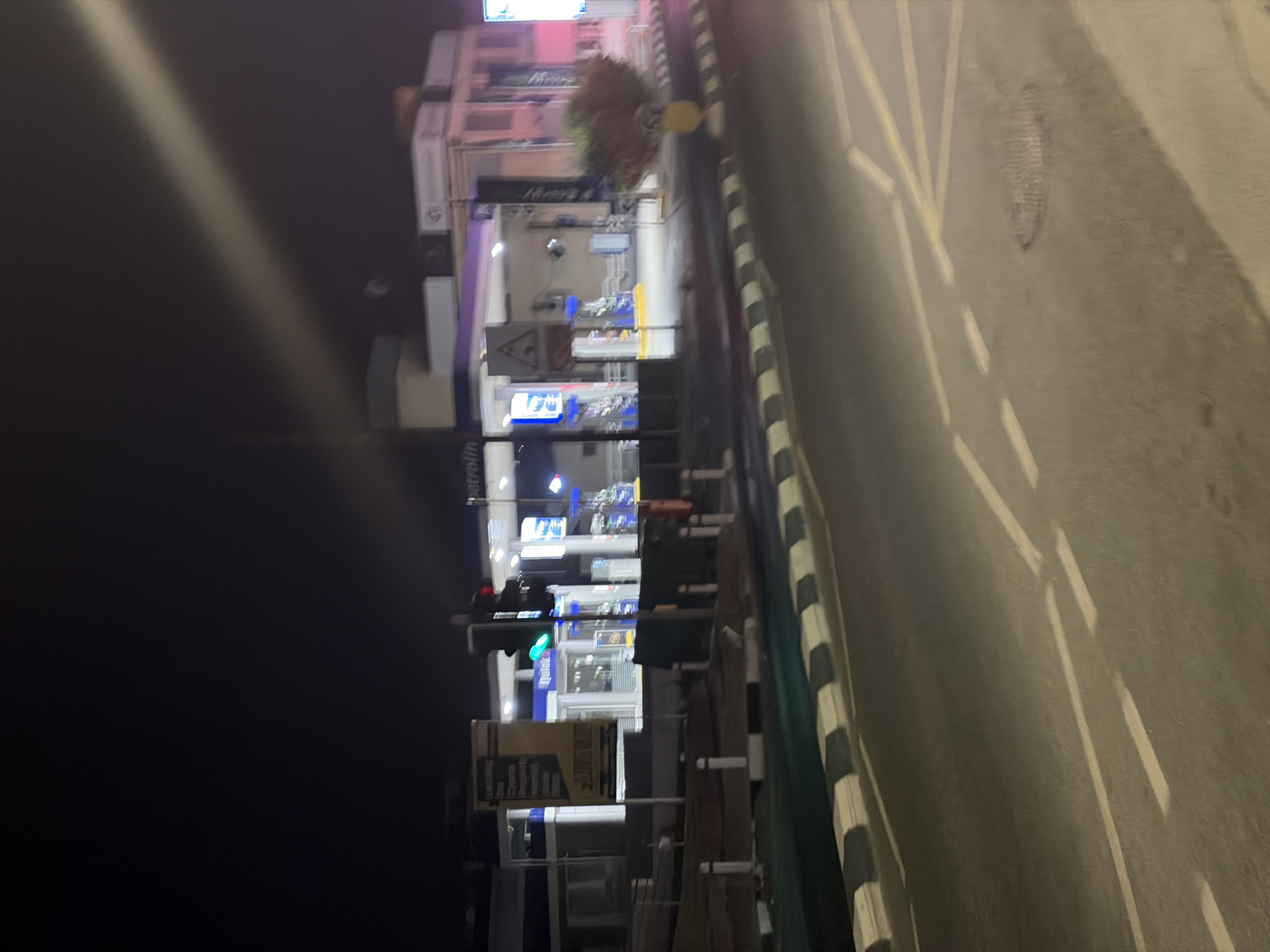 The image depicts a night scene at a petrol station. In the background, the petrol station is brightly lit with multiple fuel pumps visible. Signs and building structures are illuminated, some with blue and pink lighting. To the left of the petrol station, a large yellow sign with text is present. In the foreground, a road with white and black striped curb is visible, along with road markings. The overall scene suggests a typical urban environment at night. The context of Agios Athanasios Municipality, Cyprus is provided as the location.