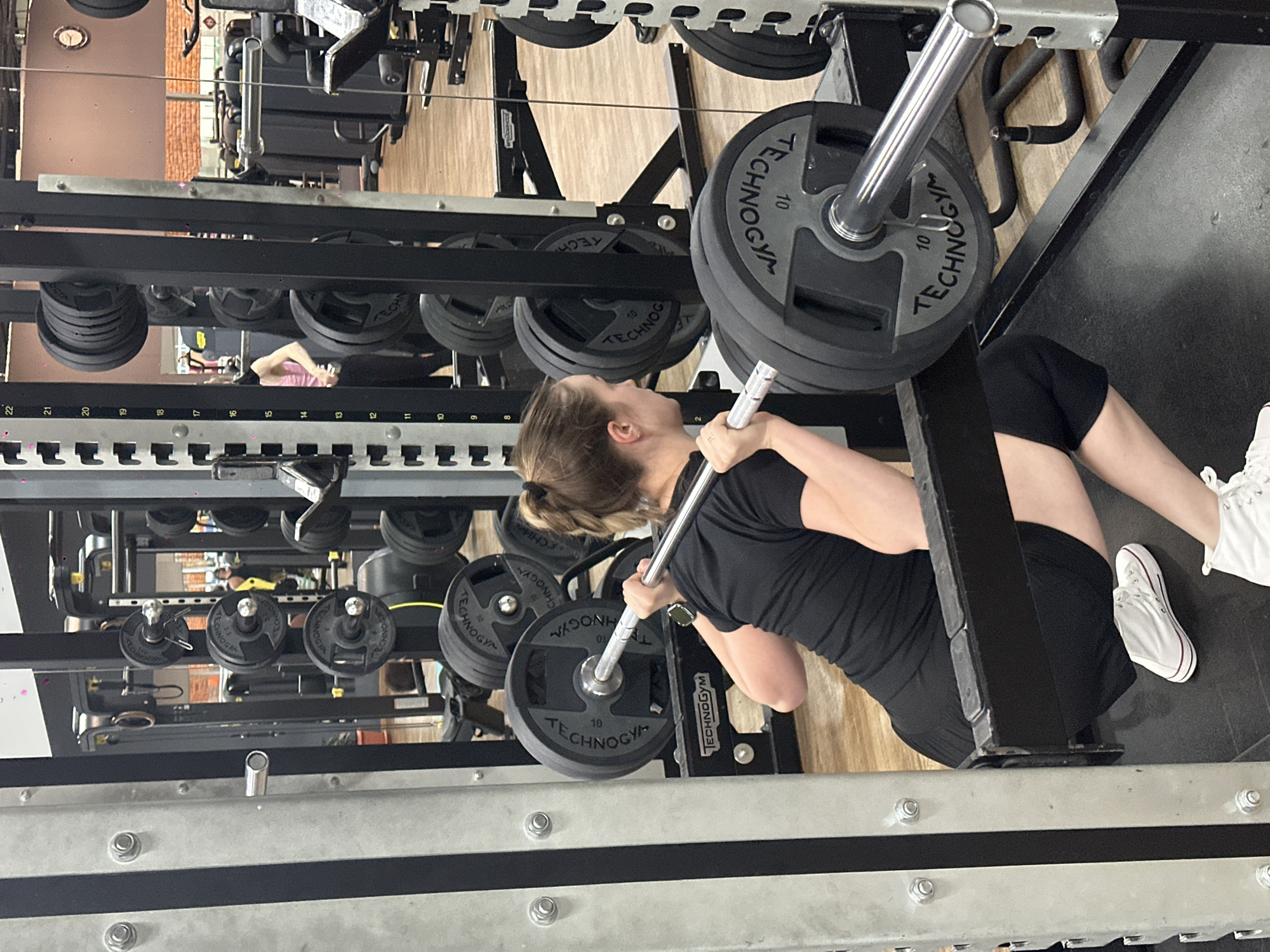 The image shows a woman performing a squat with a barbell inside a squat rack in what appears to be a gym. She is wearing a black t-shirt and shorts, with white athletic shoes. The barbell is loaded with weight plates, which are marked "TECHNOGYM" and "10". The squat rack is grey and black, with multiple weight plate holders and safety bars visible. The gym environment includes other gym equipment in the background, and a wooden floor. There is also a reflective surface, possibly a mirror, showing other people in the gym. The city of Chaikovsky, Russia is not identifiable in the image.
