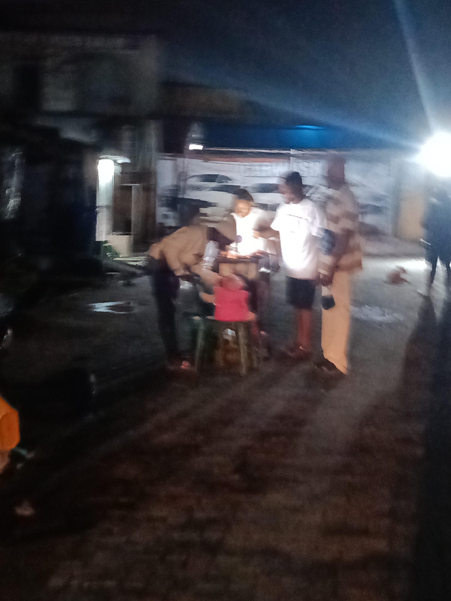 An outdoor, nighttime scene in Ado, Nigeria, features several individuals gathered around a small table with illuminated objects. The ground is wet and appears to be asphalt or concrete, with some puddles reflecting light. The background is dark and indistinct, with suggestions of structures and possibly a building with an illuminated doorway on the left.
In the foreground, a person wearing a beige top and dark pants bends over the table. To their right, a child in a pink top is seated on a stool. Another person, wearing a white t-shirt and dark shorts, stands behind the table. To their right, a person in a light-colored uniform, possibly a security guard or official, stands facing away from the camera. Another individual, wearing a dark top and pants, is partially visible on the far right, appearing to walk away.
The illuminated objects on the table suggest a possible event or activity taking place, such as a religious ceremony or a gathering. The overall impression is one of a community event happening in a dimly lit, urban environment.