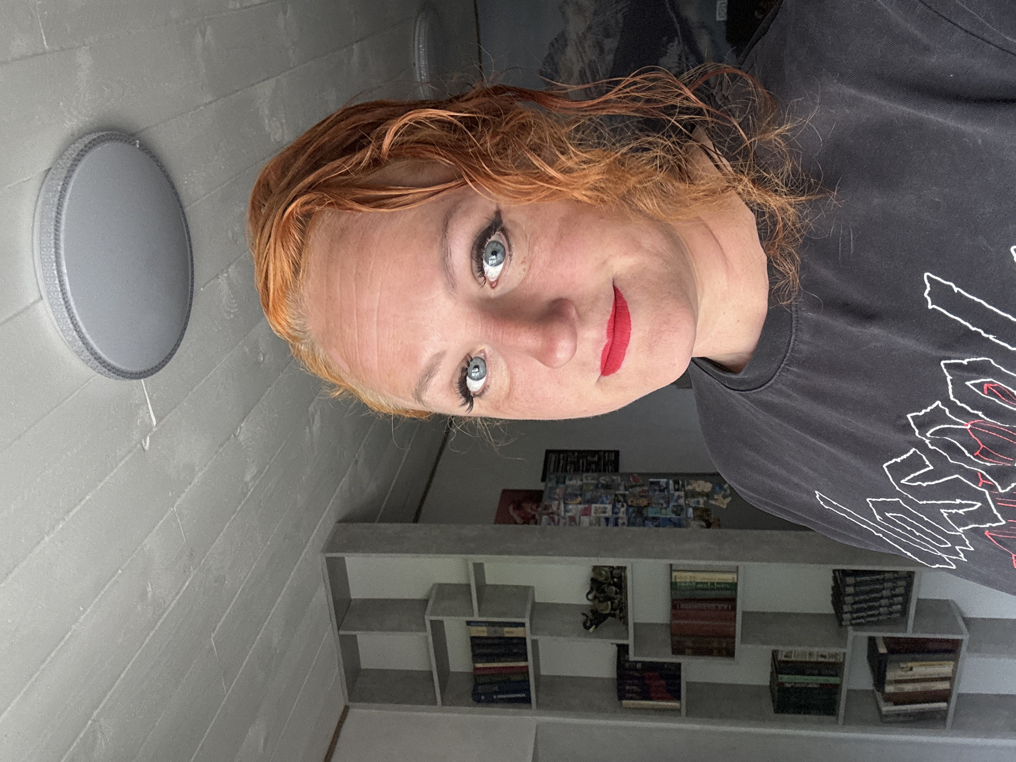 A selfie of a Caucasian woman with orange, curly hair and blue eyes is positioned in the frame. She is wearing a dark grey t-shirt with a white and red design. Her lips are painted bright red. In the background, a grey wall with horizontal planks is visible. To the left, a round grey ceiling light fixture is mounted. Behind the woman, a grey shelf unit with books and objects is present. The background also contains a collection of photographs or postcards attached to the wall. There is no specific information to suggest this image was taken in Chaikovsky, Russia.