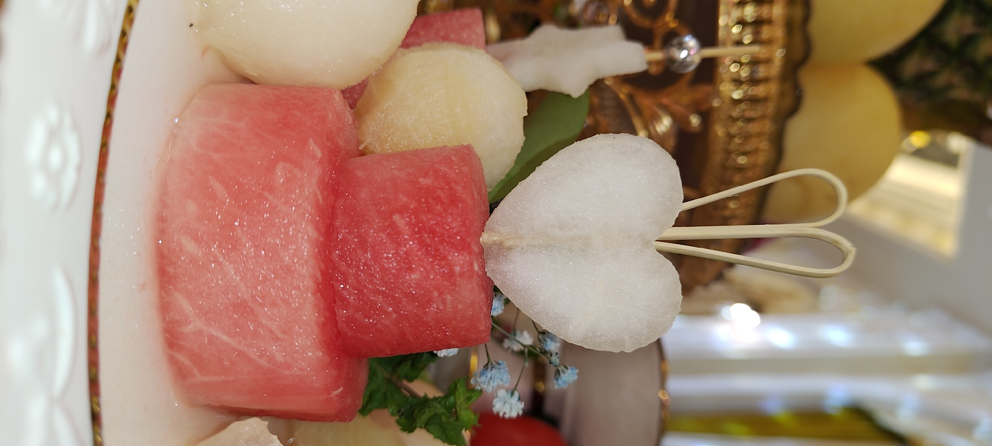 This is a close-up, eye-level, indoor, studio shot of a fruit arrangement. The arrangement is presented on a white plate with a gold rim and features a tower of watermelon cubes topped with a heart-shaped slice of what appears to be pear, impaled by a wooden skewer. The watermelon cubes are of varying sizes and are stacked in two layers, with the bottom layer being larger than the top. To the left of the watermelon tower, there are spherical pieces of what looks like melon or pear. In the background, out of focus, is a golden tray holding more fruit, possibly melons, and a white ornate bowl with a gold rim. There are also blurred elements of soft lighting and what appear to be decorative white fabric drapes. Small blue flowers and green leaves are visible to the right of the watermelon tower, adding a touch of garnish. The scene is well-lit, with the focus sharp on the fruit arrangement in the foreground, creating a shallow depth of field that blurs the background. The overall impression is one of a carefully prepared dessert or appetizer, possibly for a celebration or a restaurant setting. This image is not specific to the city of Khujand, Tajikistan, as there are no identifiable landmarks or contextual clues that would place it there.