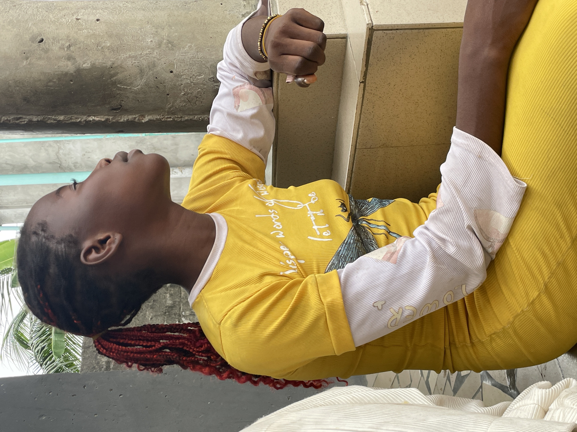 A young person with dark skin and prominent red braided hair extensions is captured in a close-up, portrait-style image, gazing upwards with a calm and contemplative expression. They are dressed in a vibrant yellow ribbed long-sleeve shirt, under which a white long-sleeve undershirt is visible at the collar and cuffs. A golden beaded bracelet adorns their left wrist.
The yellow shirt features white text, partially readable as "Wisdom Words of..." and a graphic design that appears to be a dragonfly or similar insect. The white undershirt sleeves also show faint patterns, possibly depicting flowers or figures, along with small clover-like shapes and text that includes the word "POWER." The person's lower body is clad in a matching yellow ribbed garment, likely a skirt or pants.
The background suggests an outdoor or semi-outdoor setting. A textured concrete surface is visible above and to the left of the subject, while a tiled wall is seen to the right. Green foliage, specifically palm leaves, can be observed on the far left, indicative of a tropical or subtropical environment like Badagry, Nigeria. In the bottom foreground, a white ribbed fabric, possibly a cushion or garment, is partially visible. The scene is brightly lit, suggesting it is daytime.