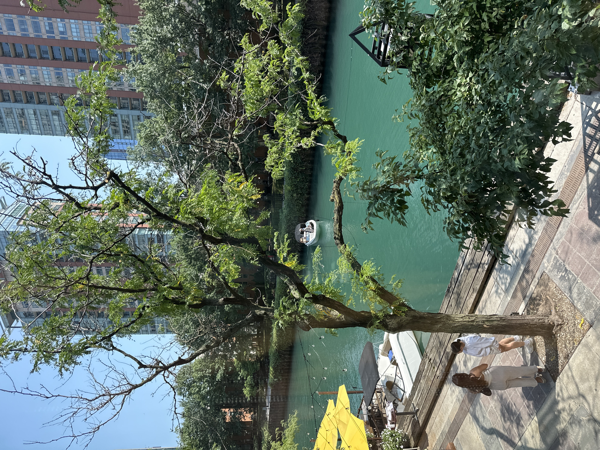 This is an image of the Chicago River in Chicago, Illinois. Two women are standing on a sidewalk next to the river. One woman is wearing a white top and striped pants, and the other is wearing a white sweatshirt and shorts. They are facing each other and talking. A boat is docked on the river, and a few other boats are in the distance. There are trees and buildings along the riverbank. The sun is shining, and the sky is blue.