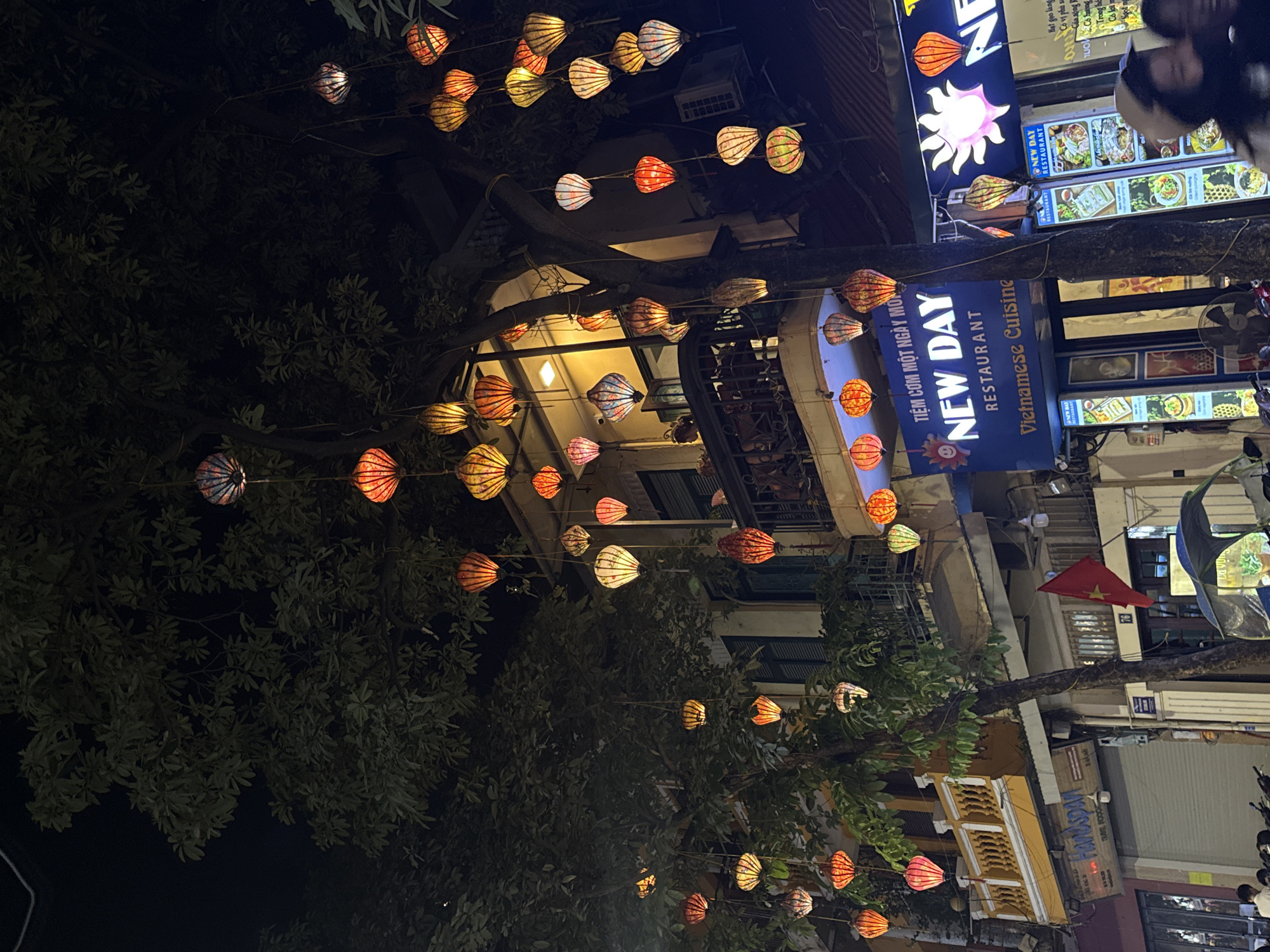 The image shows a street scene at night in Hà Nội, Vietnam. Lanterns hang overhead, strung between trees and buildings, creating a festive atmosphere. Signs for "New Day Restaurant" are prominently displayed. The signs indicate "Tiệm cơm một ngày mới," translating to "A New Day Restaurant." The restaurant serves Vietnamese cuisine. A Vietnamese flag is visible, and there are nearby buildings with architectural features typical of the area. Other signs are visible but not clearly legible.