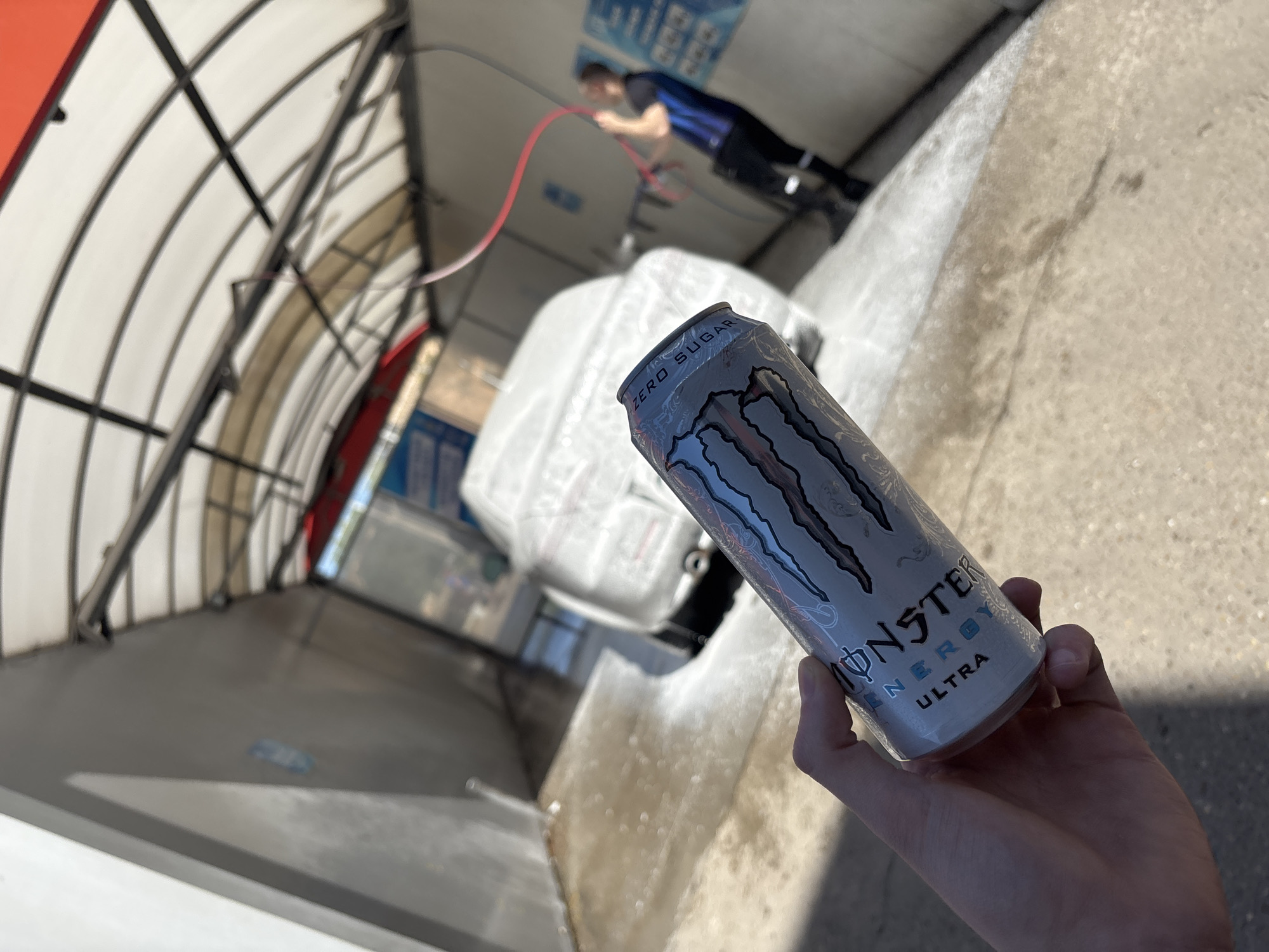 A person is holding a can of Monster Energy Ultra in their right hand at a car wash in Ruse, Bulgaria. A man in a dark blue and black shirt and dark pants is visible in the background, operating a red hose. The car wash appears to be a drive-through type, with metal structures overhead. A white car is positioned in the wash bay. The ground is a concrete surface.