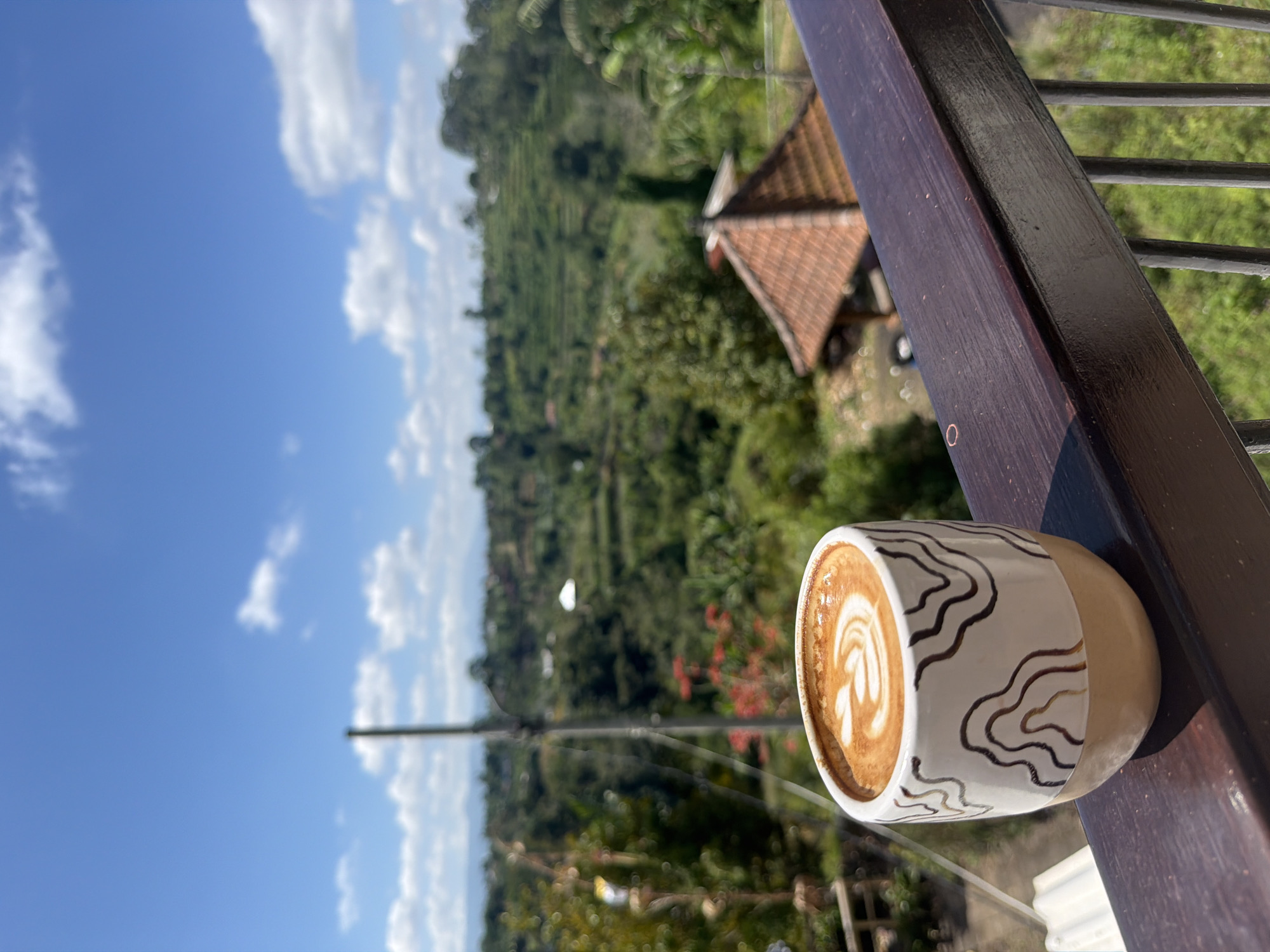 A cup of coffee with latte art sits on a dark wooden railing overlooking a lush green landscape in Baturiti, Indonesia. The coffee is in a white ceramic cup with a tan base, decorated with thin, wavy brown lines. In the background, rolling hills covered in vegetation descend towards a village nestled among the trees. A traditional Balinese-style building with a terracotta roof is visible in the mid-ground. The sky is a vibrant blue with scattered white clouds. The scene suggests a tranquil setting, possibly a cafe or a private residence with a scenic view.