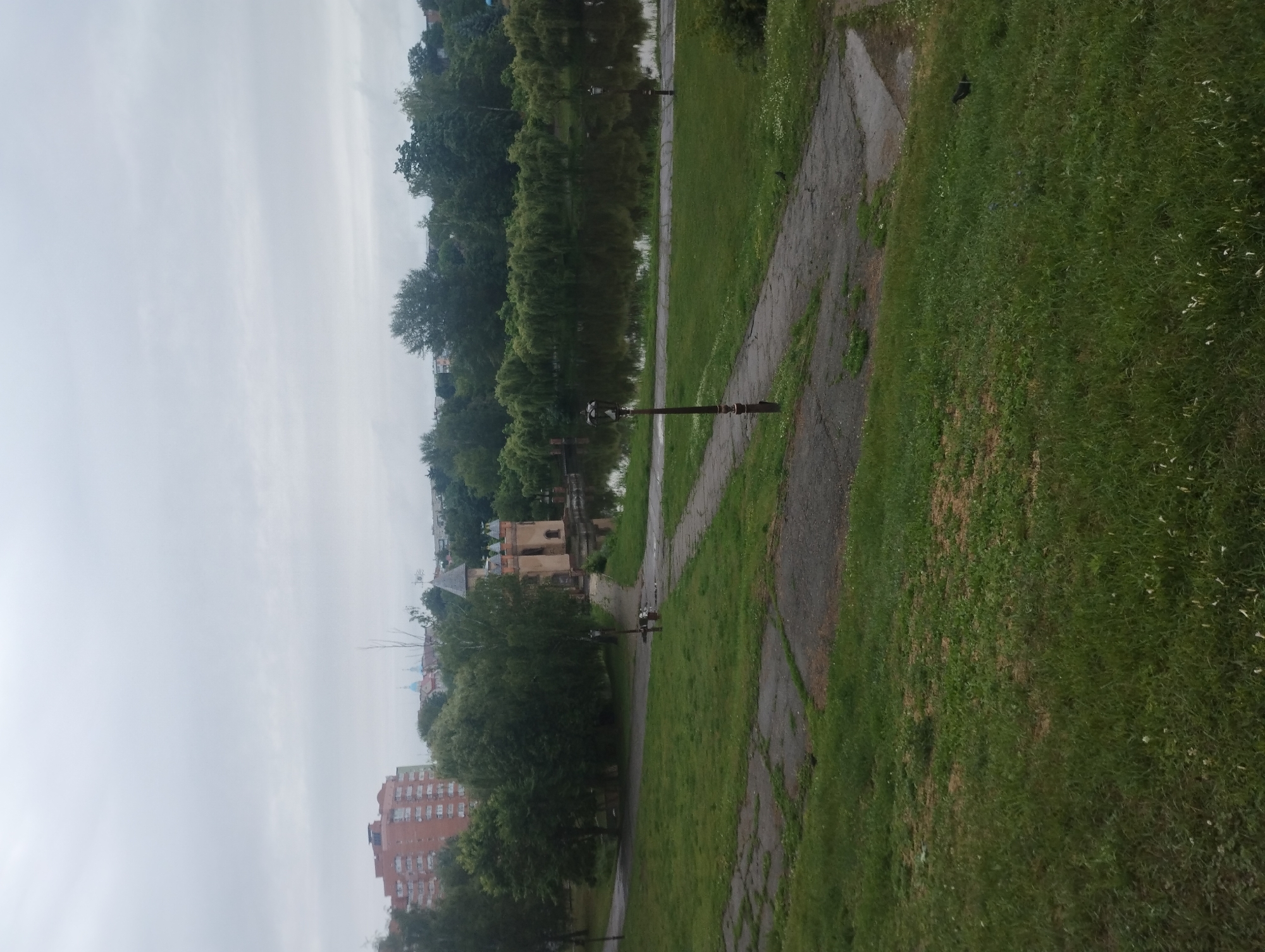 The image depicts a tranquil park scene in Sumy, Ukraine, likely on an overcast day. In the background, a distinctive red brick apartment building stands tall, a common sight in urban areas. Further in the distance, the blue domes of a church are visible, adding a touch of spiritual architecture to the cityscape.
The foreground is dominated by a lush green lawn, interspersed with patches of grey, cracked asphalt paths that wind through the park. A few trees dot the landscape, their leaves appearing a vibrant green. In the middle ground, a serene body of water reflects the overcast sky and the surrounding trees. On the water's edge, a quaint, castle-like building with a pointed roof and crenellated turrets stands out. The building's facade is a pale ochre, hinting at its age and possibly historical significance.
Several lampposts are strategically placed along the paths, their ornate designs suggesting a classic park aesthetic. A couple of individuals are seated on a bench near the water, their presence adding a sense of human activity to the otherwise peaceful environment. The overall impression is one of a well-maintained public space, offering a respite from the urban bustle.