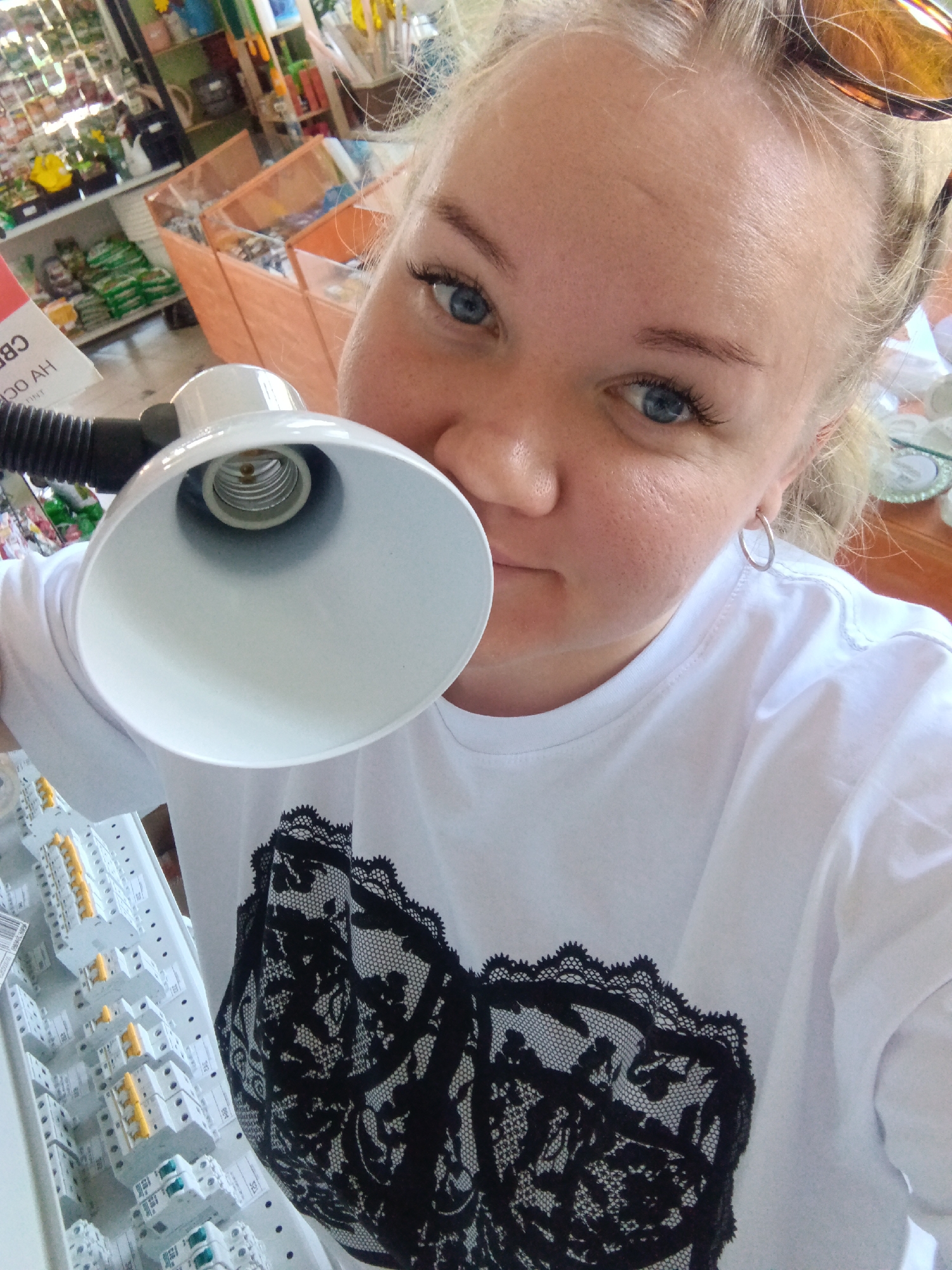 A fair-skinned woman with blonde hair pulled back is holding a white lamp fixture close to her face. She is wearing a white t-shirt with a black lace design on the front. The lamp is held at an angle, revealing its interior and a black flexible neck. The woman has blue eyes and is wearing hoop earrings. Sunglasses are perched atop her head.
To the left of the frame, a shelf displays electrical components, including white and yellow units. In the background, a store setting is visible with shelves stocked with various goods, including gardening supplies and other merchandise. A sign with Cyrillic writing can be seen partially visible in the background.