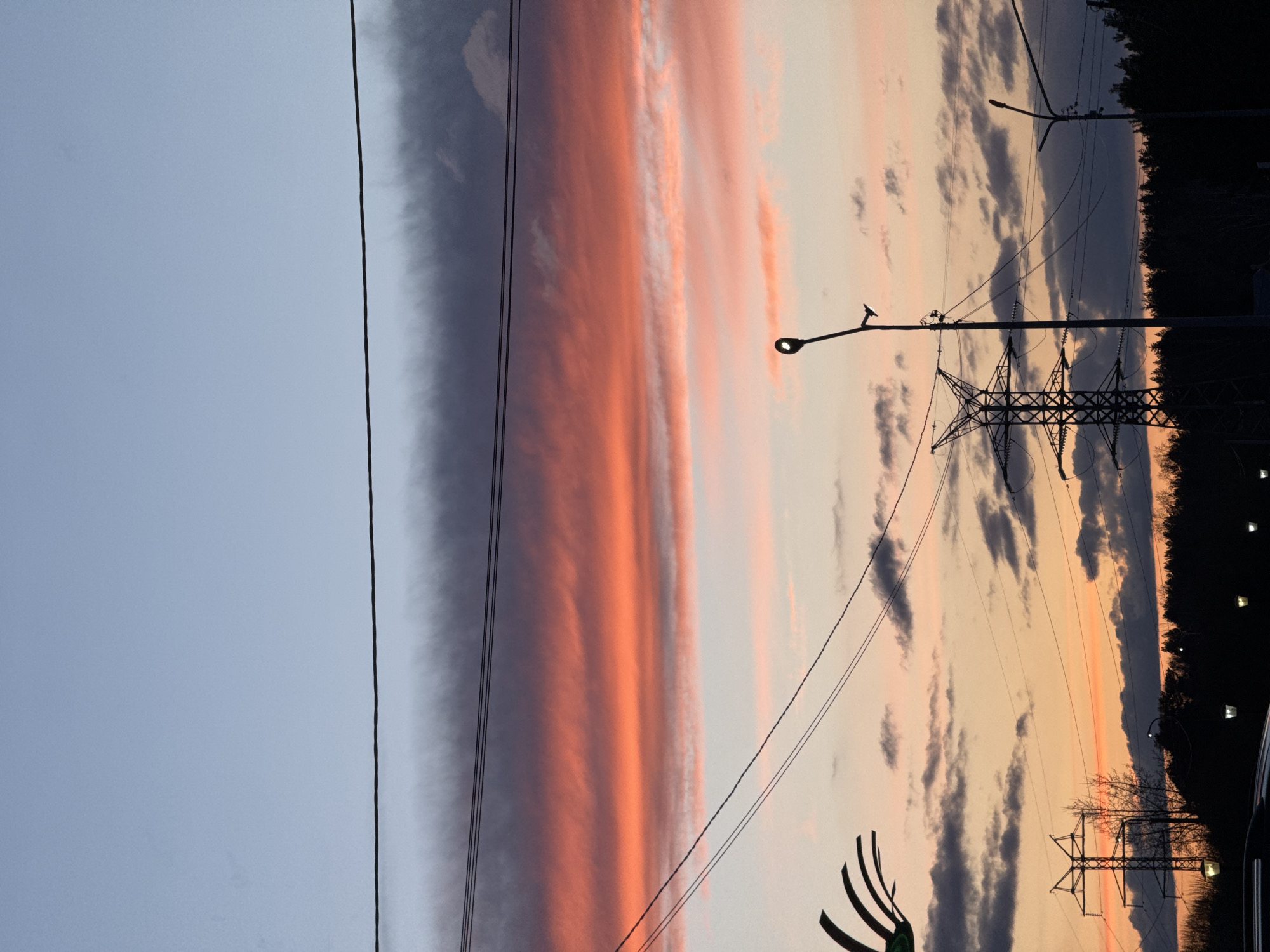 The image depicts an outdoor scene at either sunrise or sunset, characterized by a dramatic sky over what appears to be a rural or suburban landscape in Chaikovsky, Russia. The sky dominates the frame, displaying a gradient from a pale blue-grey at the top to vibrant red and orange hues across the middle, forming a prominent, elongated horizontal cloud band. Below this, the sky transitions to softer pinks and pale oranges, with scattered darker grey clouds.
Silhouetted against the colorful sky are various elements. Multiple power lines extend horizontally across the scene, supported by several utility poles and a large, imposing electricity pylon. Several streetlights are visible, some appearing illuminated, providing small points of light against the fading daylight. Along the horizon, a dark, dense silhouette of a forest or tree line stretches across the bottom portion of the image. A small, dark section of a vehicle is partially visible on the far right edge, and an abstract, dark, claw-like or fan-like object is present in the bottom left corner.
The time of day is clearly indicated by the warm, intense colors of the clouds and the contrasting dark silhouettes of the foreground objects and horizon. No specific human activity or visible text is present.
