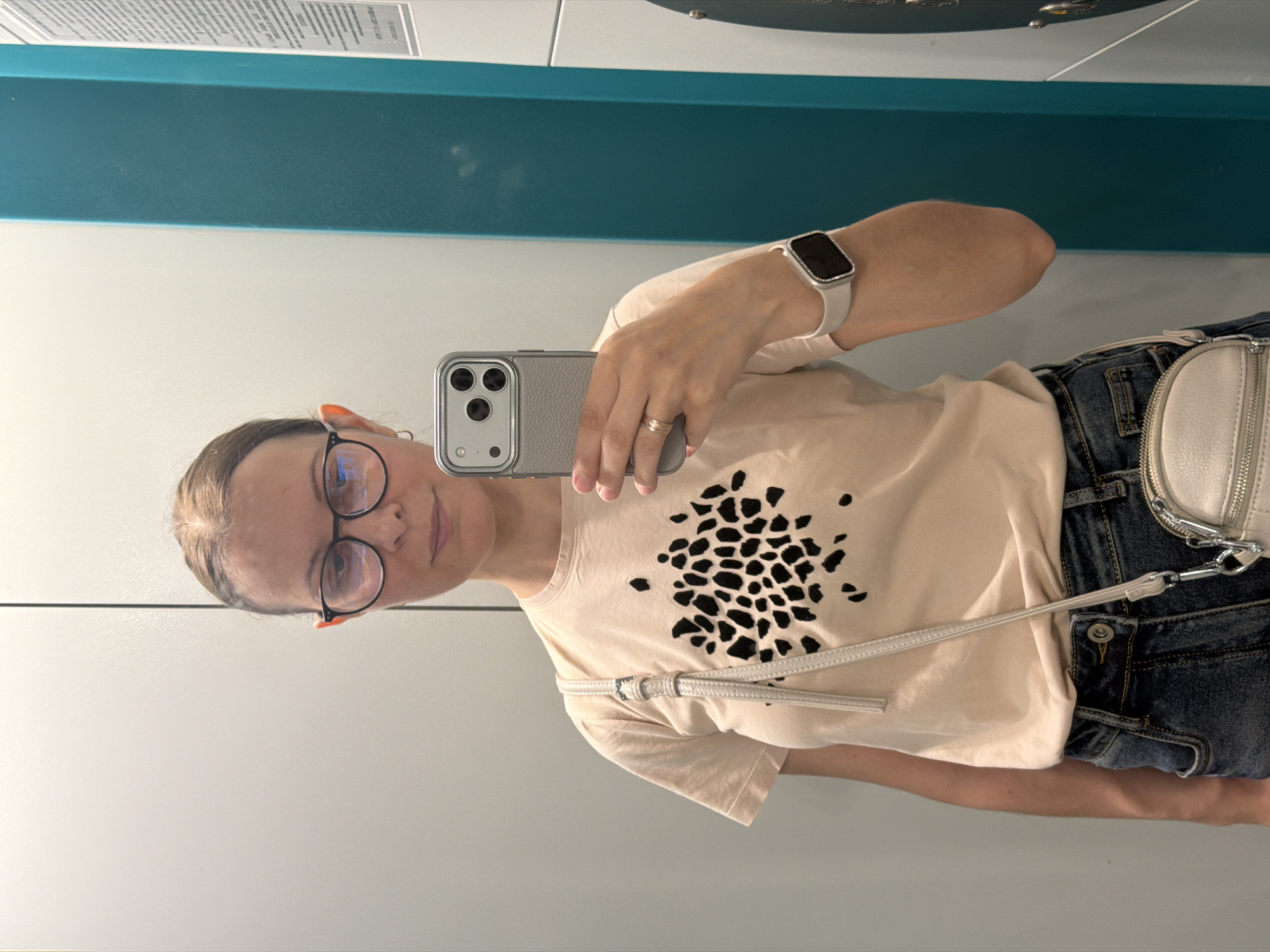 A woman is taking a mirror selfie in a public restroom, possibly in Izhevsk, Russia. She is wearing glasses, a light-colored t-shirt with a black abstract print, blue jeans, and a white crossbody bag. She has a smartwatch on her left wrist and a ring on her right ring finger. The background consists of a white wall, a teal-colored partition, and a sliver of what appears to be a washing machine or dryer.
