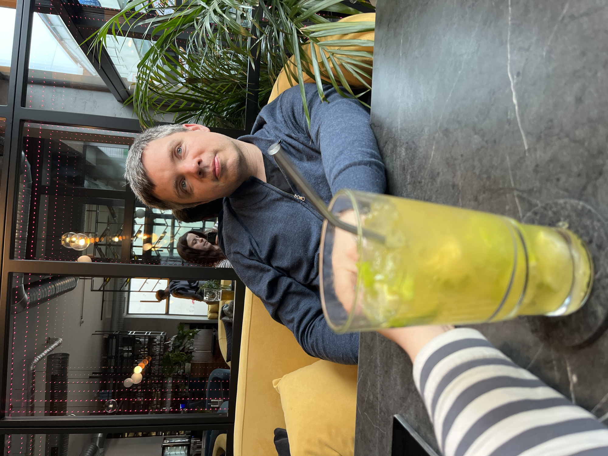 The image shows a man sitting at a dark-colored table inside what appears to be a restaurant or bar, based on the interior design. He is wearing a blue hooded jacket and has short, graying hair. In front of him is a tall glass containing a yellow-green beverage with ice. A person wearing a striped gray and white shirt is holding the glass. In the background, there is a glimpse of another person sitting on a yellow sofa. The restaurant has a modern design with exposed piping and a large window. The location is Bad Kreuznach, Germany.