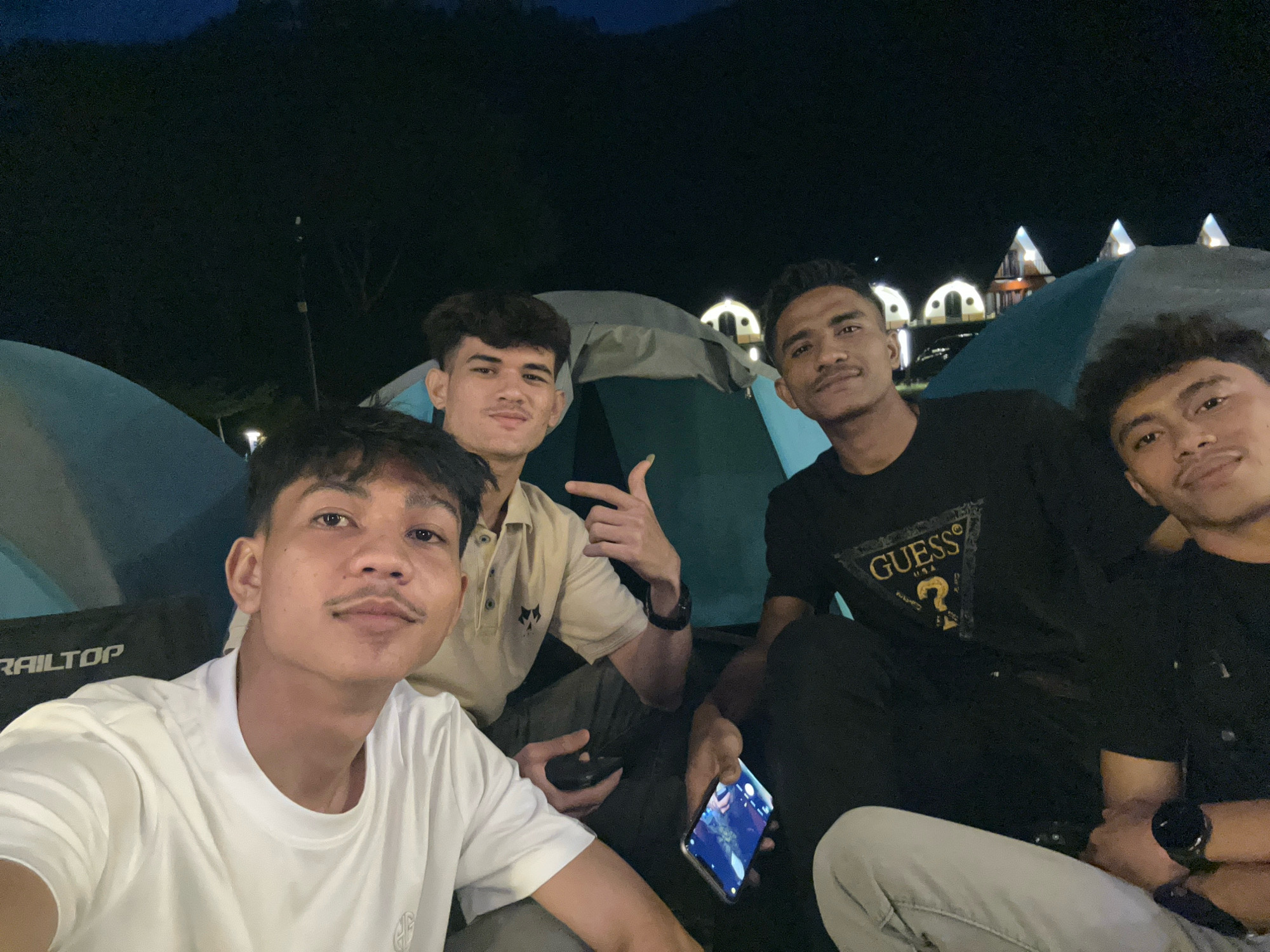 Four young men are posing for a selfie at night, likely at a campsite in Merodot, Indonesia, given the context. The man in the foreground on the left wears a white t-shirt and has a mustache. To his right, a man in a tan polo shirt with a dark emblem on the chest is making a peace sign. Behind them, a man in a black t-shirt with a "GUESS USA" logo is looking towards the camera, and to his right, another man in a black t-shirt is partially visible. They are seated in front of tents, and in the background, illuminated structures resembling dome-shaped cabins are visible, suggesting a glamping or resort setting. The overall scene is dimly lit by artificial lights and the night sky.